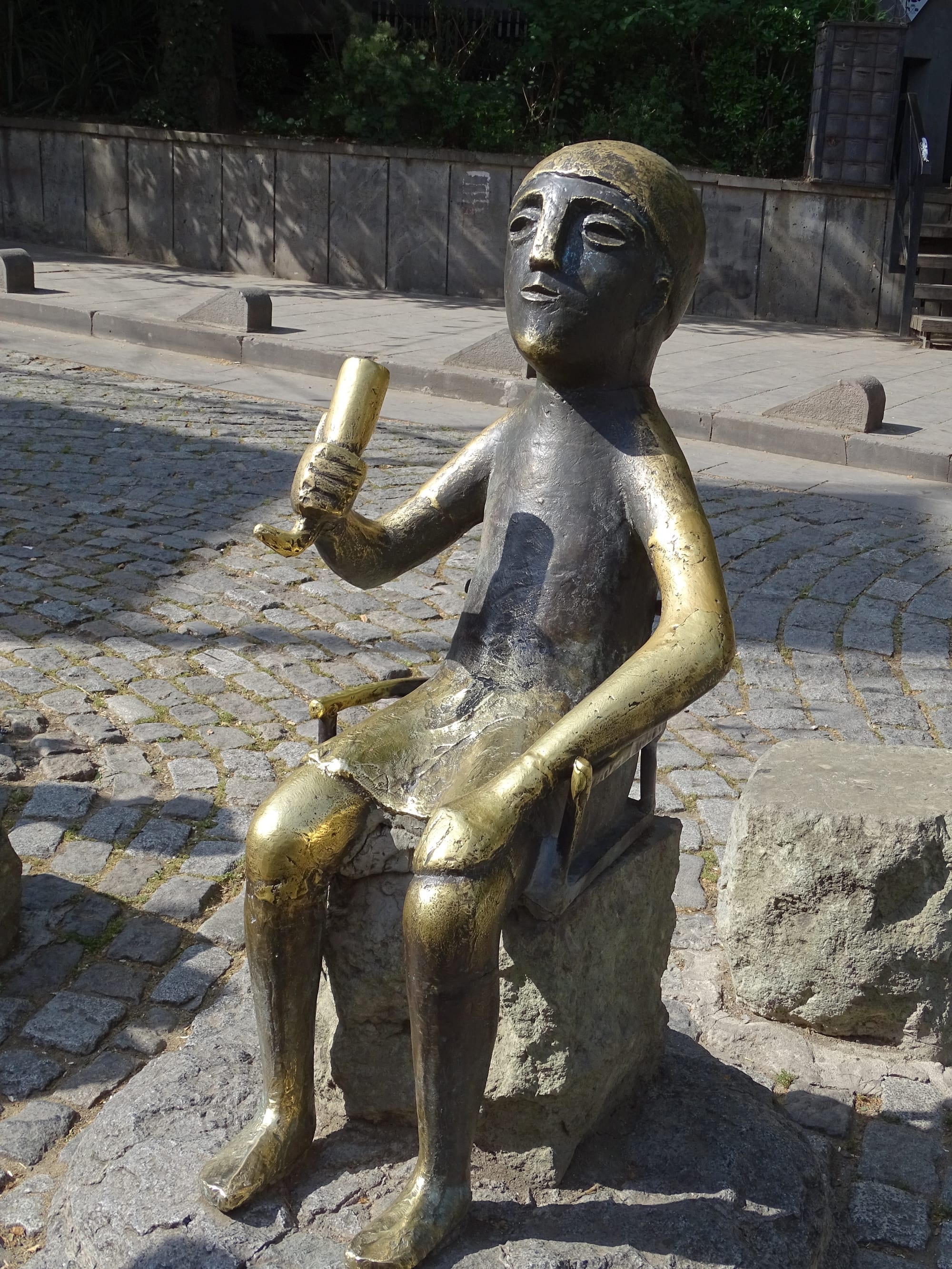 A large bronze statue of a seated man with a simple mask-like face, raising a drinking horn in his right hand, set on a stone block on a cobbled street
