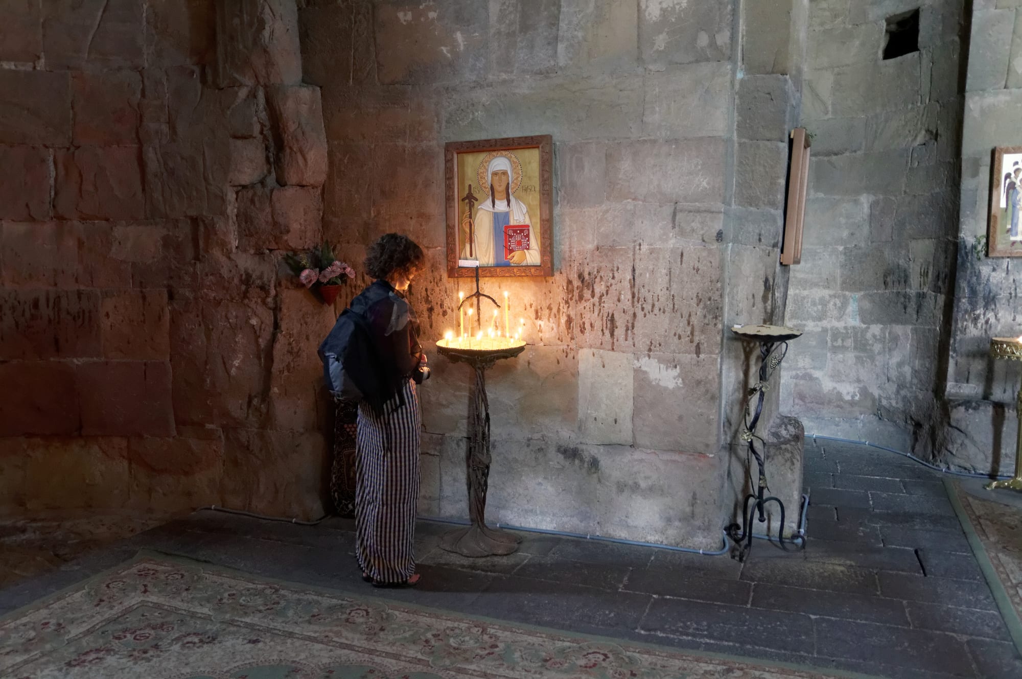 Thin lit candles standing in trays of sand on two stands inside a bare stone church, with a painted icon on the wall and a woman seen from behind near a wooden cross