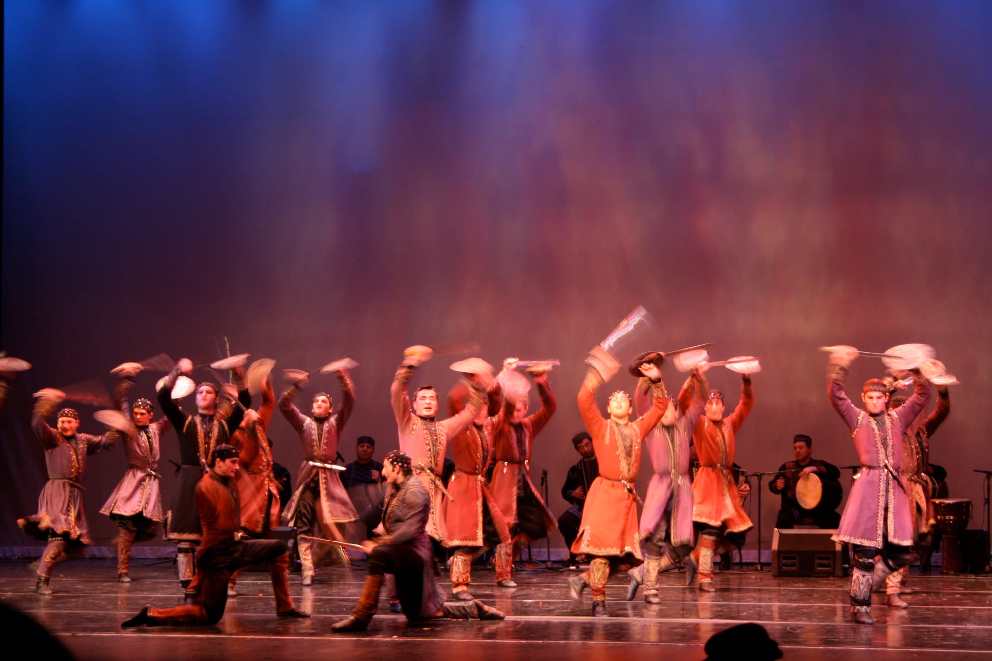 A line of male dancers in long red, pink and purple coats leaping on a stage and swinging their hats above their heads, with two dancers kneeling in front and musicians with drums at the back