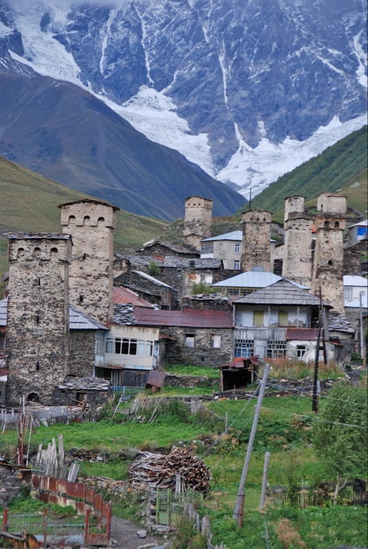 A village of stone defensive towers on a green valley floor below snow peaks