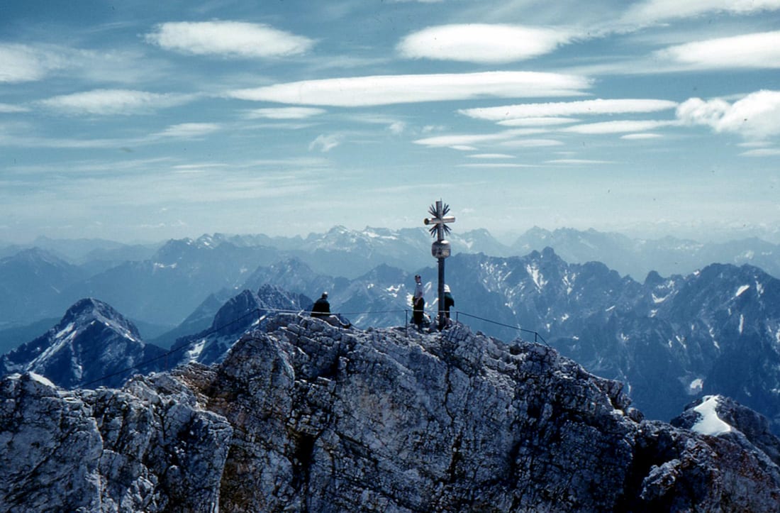 A gilded cross on a rocky summit crowded with visitors, snow peaks all around