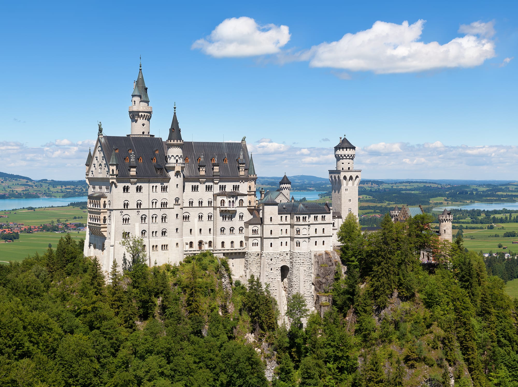 A white fairy-tale castle with towers and turrets on a forested crag below mountains