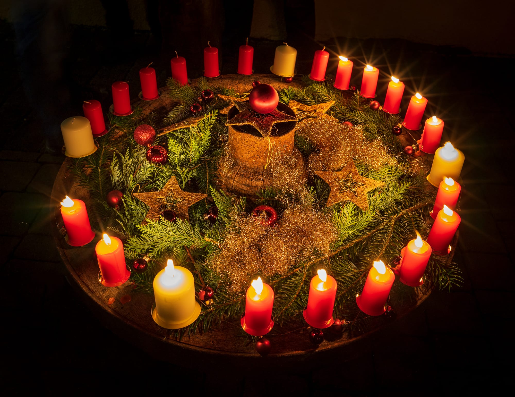 A large round wreath of fir branches in the dark ringed by lit candles, small red ones for weekdays and larger white ones for Sundays, decorated with red baubles and golden stars