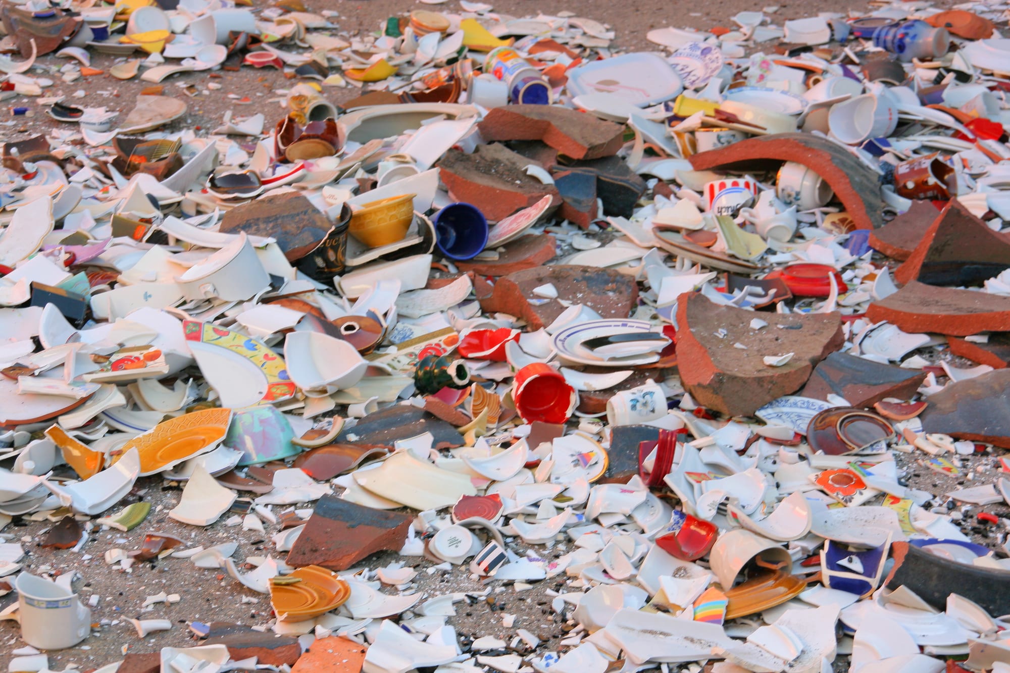 A thick layer of broken china cups, plates and bowls in white and bright colours scattered over the ground, mixed with pieces of terracotta