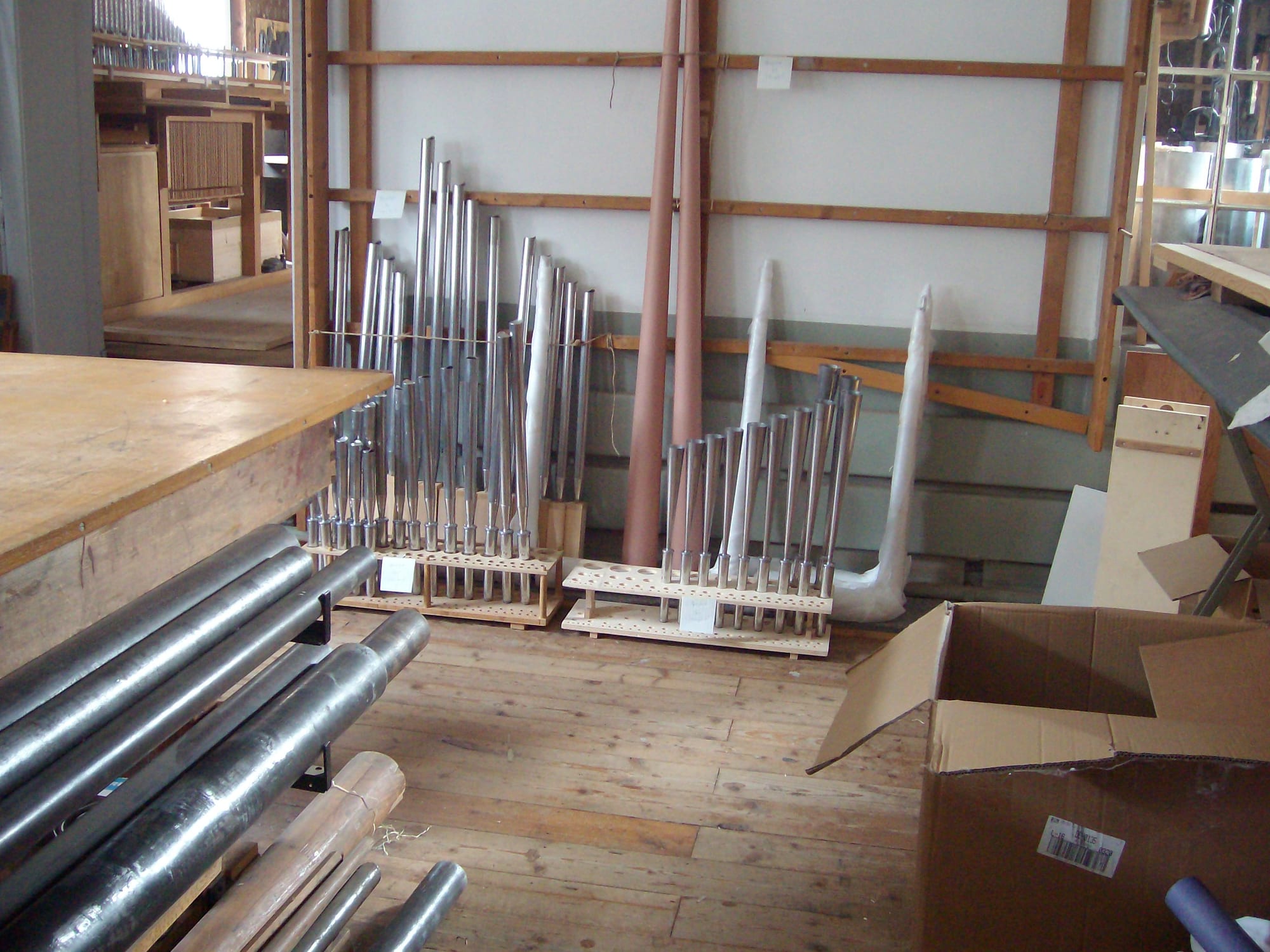 Rows of shiny metal organ pipes of different lengths standing upright in wooden racks in a workshop with a plank floor, with long tubes lying on a rack in the foreground