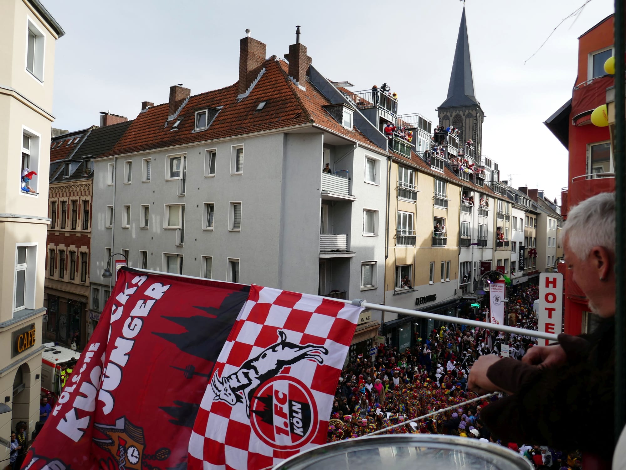 A narrow street between tall city houses packed with a crowd in colourful costume, seen from a balcony draped with red and white flags