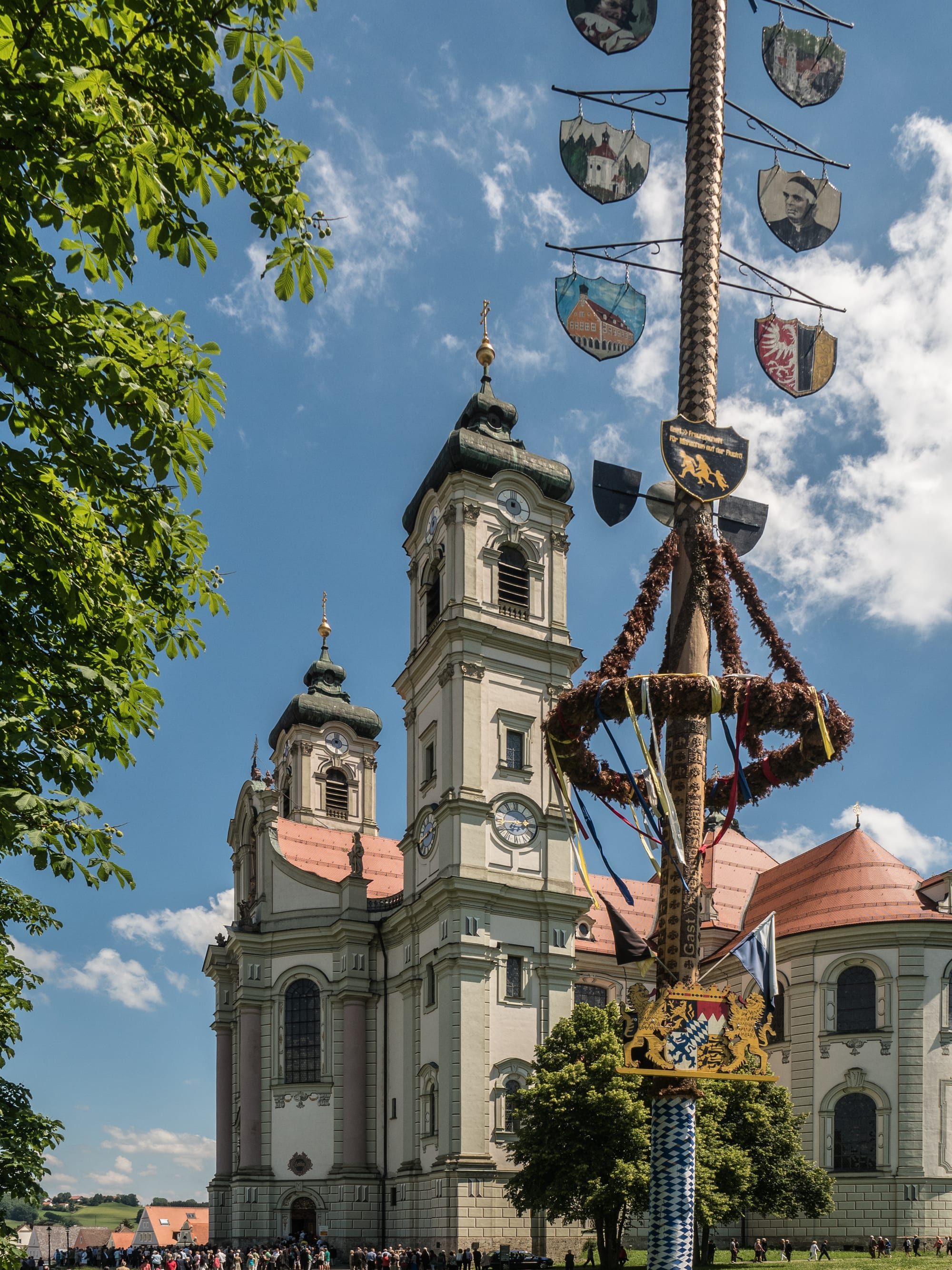 A tall maypole hung with painted shields, a green garland and ribbons stands in front of a twin-towered white baroque basilica under a blue sky