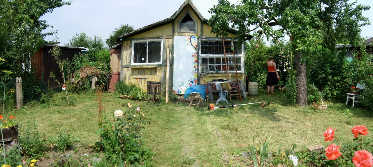 A small painted garden shed surrounded by flowerbeds, fruit trees and a mown lawn, with a woman standing among the plants