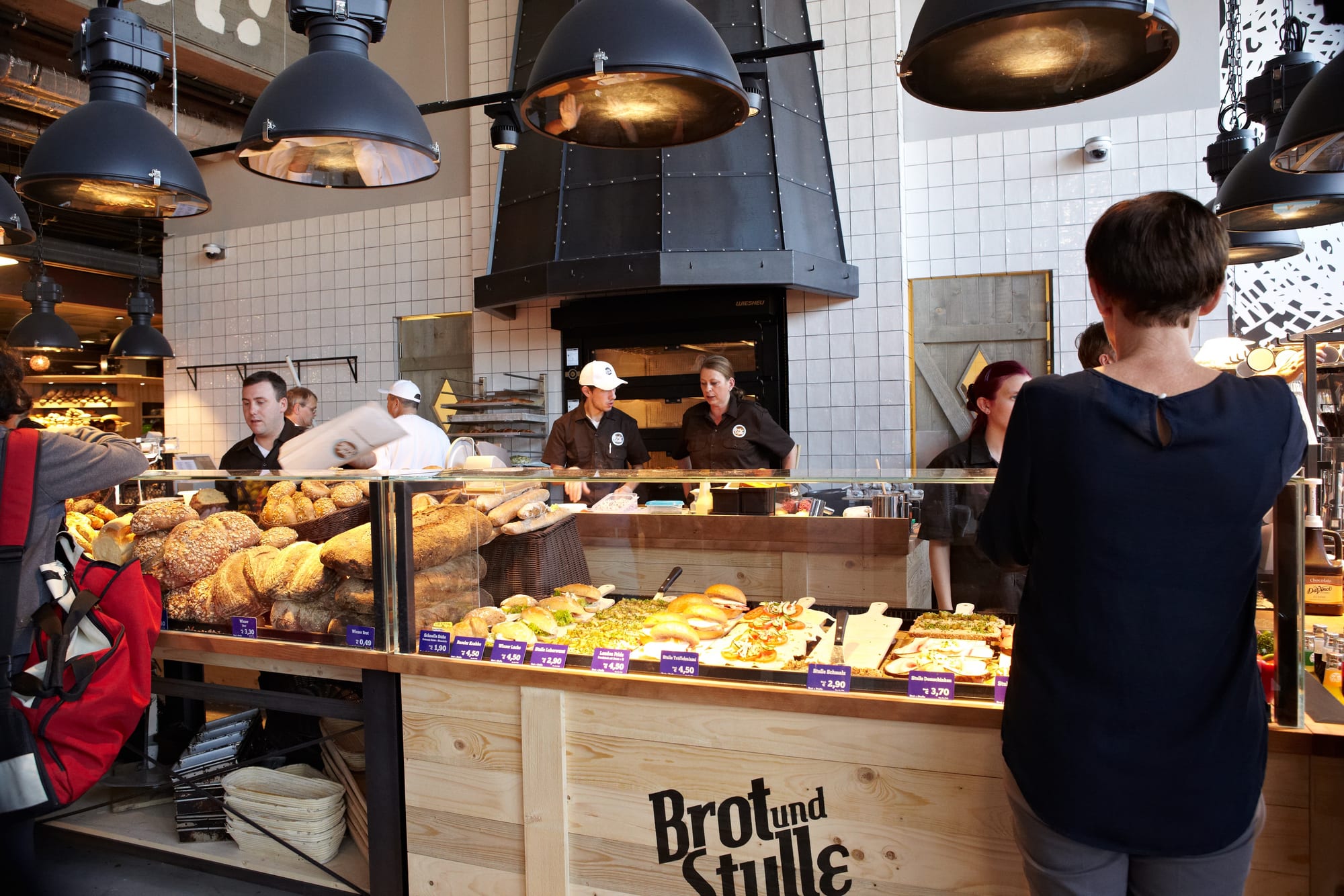 A bakery counter piled with loaves and filled rolls, with staff working behind it and a customer in front