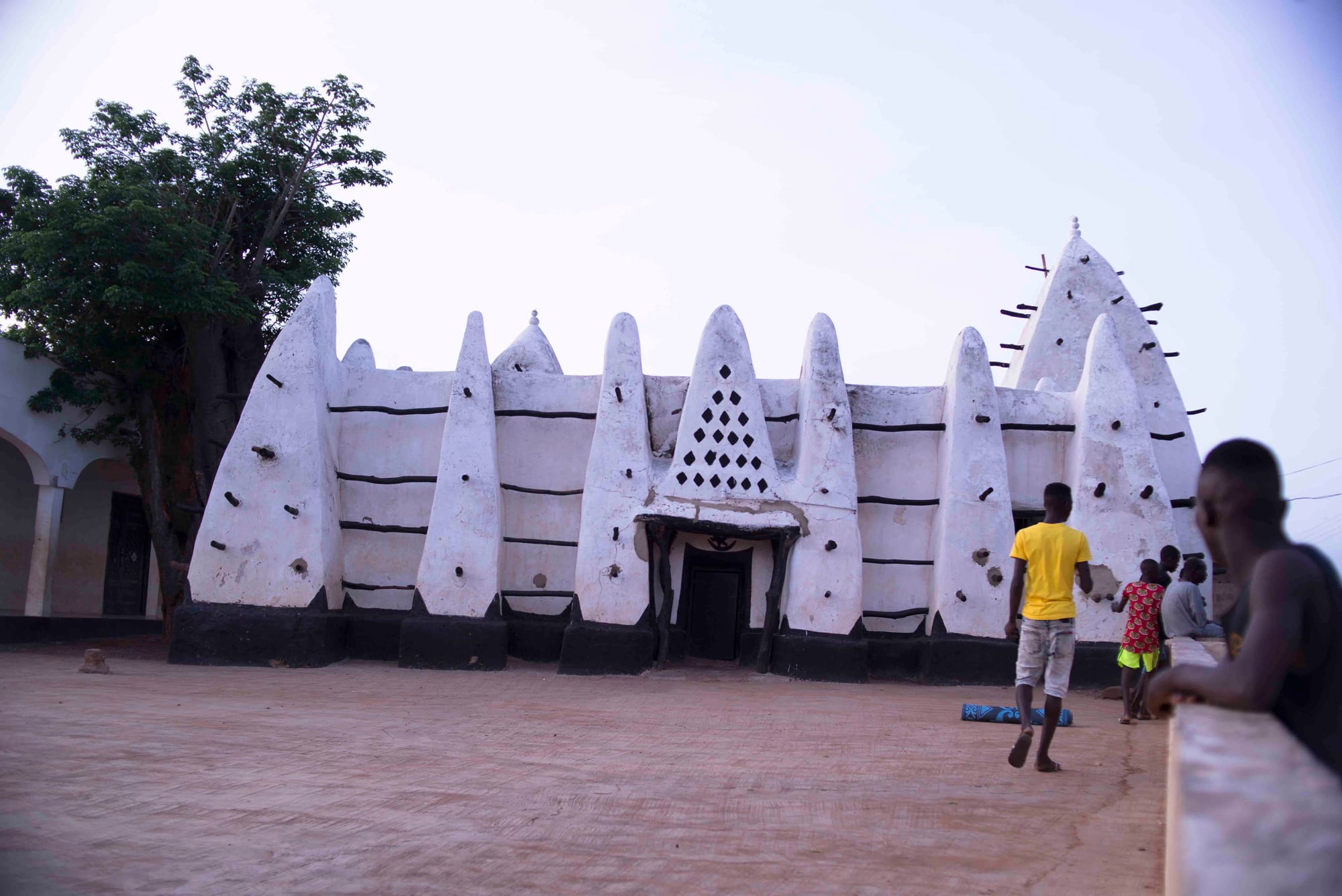 A low whitewashed mud-built mosque with tall pointed buttresses and a tapering tower, studded with protruding wooden beams, beside a tree, with a few people walking in the sandy square in front