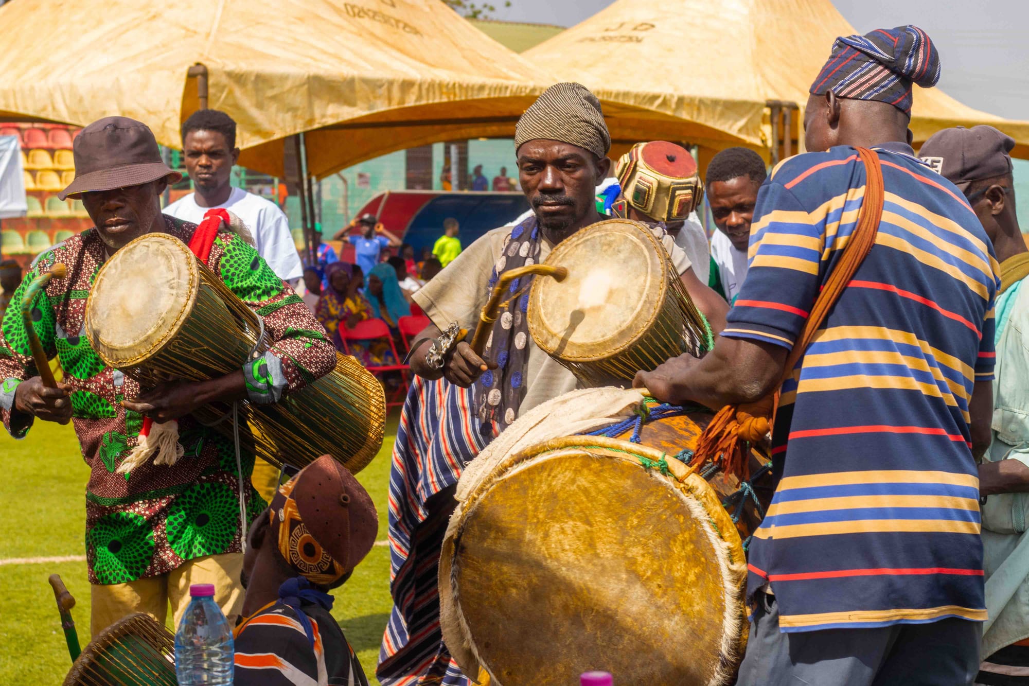 Several men in patterned smocks and caps play large hourglass-shaped and barrel drums with curved sticks at an outdoor festival, with tents and spectators behind