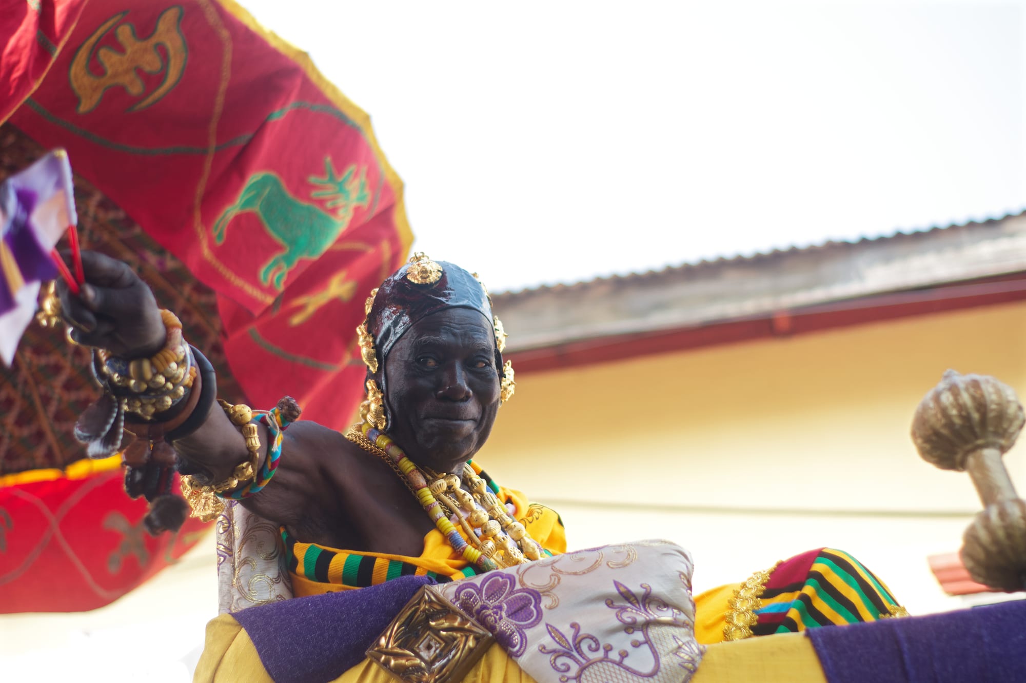 An elderly chief wearing gold jewellery, beads and bright kente cloth smiles and raises a hand from a palanquin beneath a red ceremonial umbrella