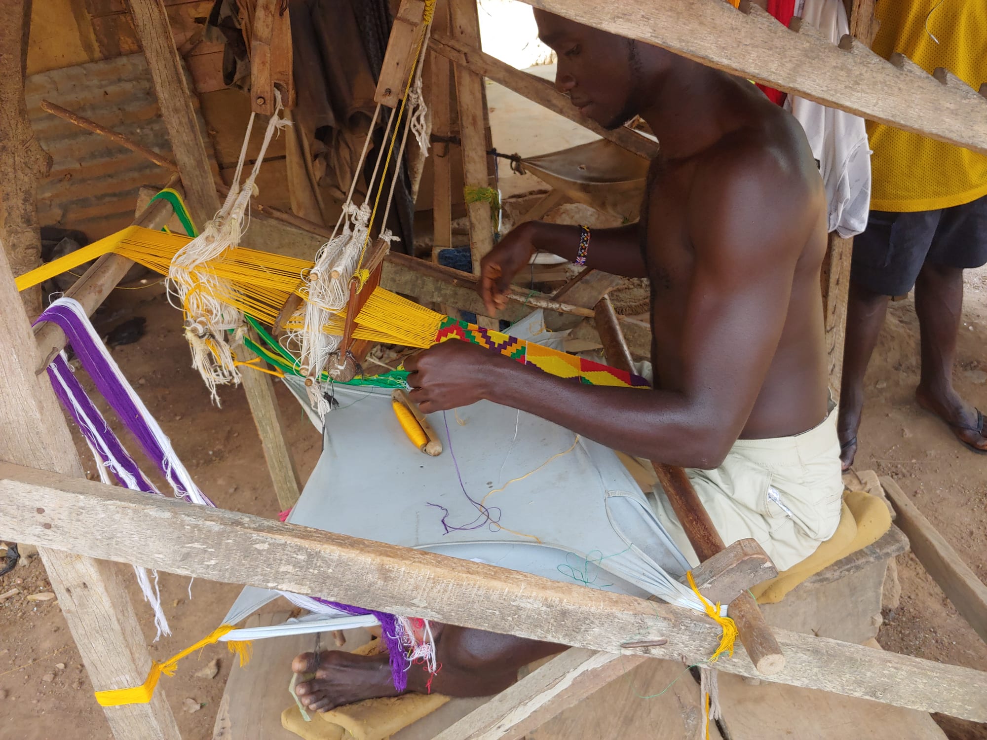 A weaver sits at a wooden loom working a narrow strip of kente cloth in yellow, green, red and purple, with bundles of bright yellow thread stretched out in front of him
