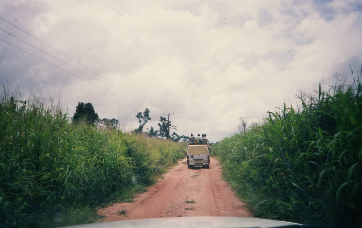An overloaded tro-tro minibus with passengers riding on the roof, driving down a red dirt road through tall grass