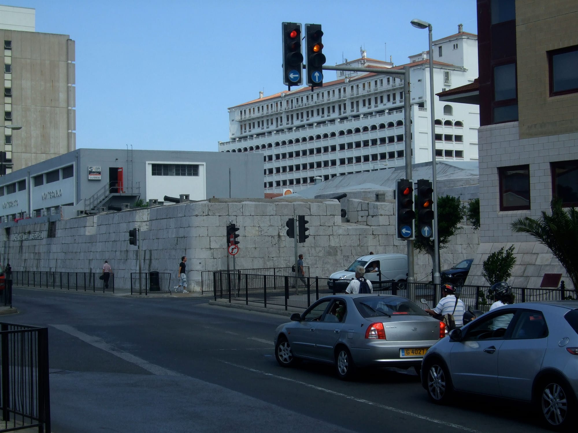 A city street with cars stopped at traffic lights beside an old stone fortress wall and modern buildings