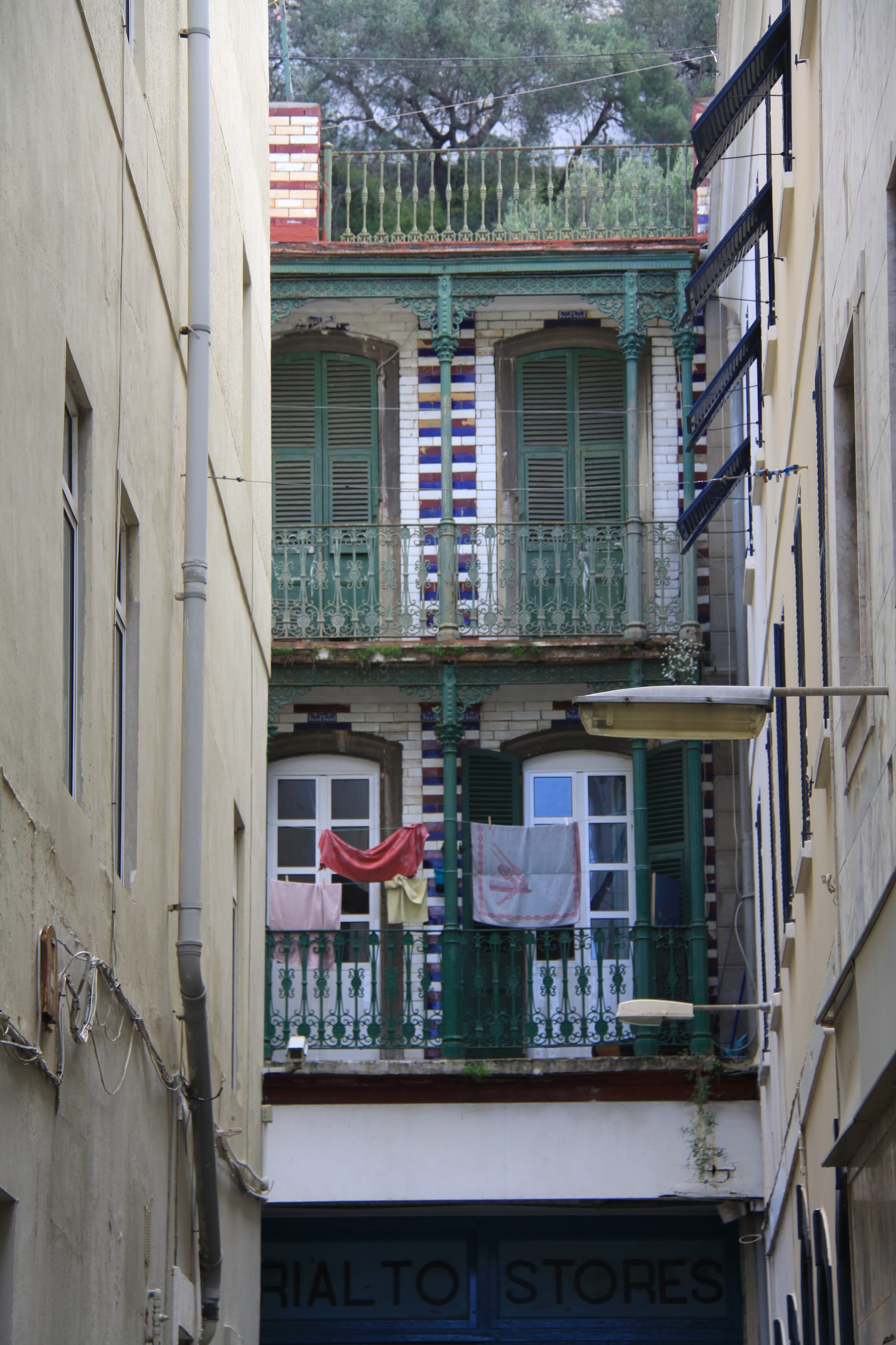 A narrow gap between tall buildings in Gibraltar, with ornate balconies, shuttered windows, and laundry hanging out to dry