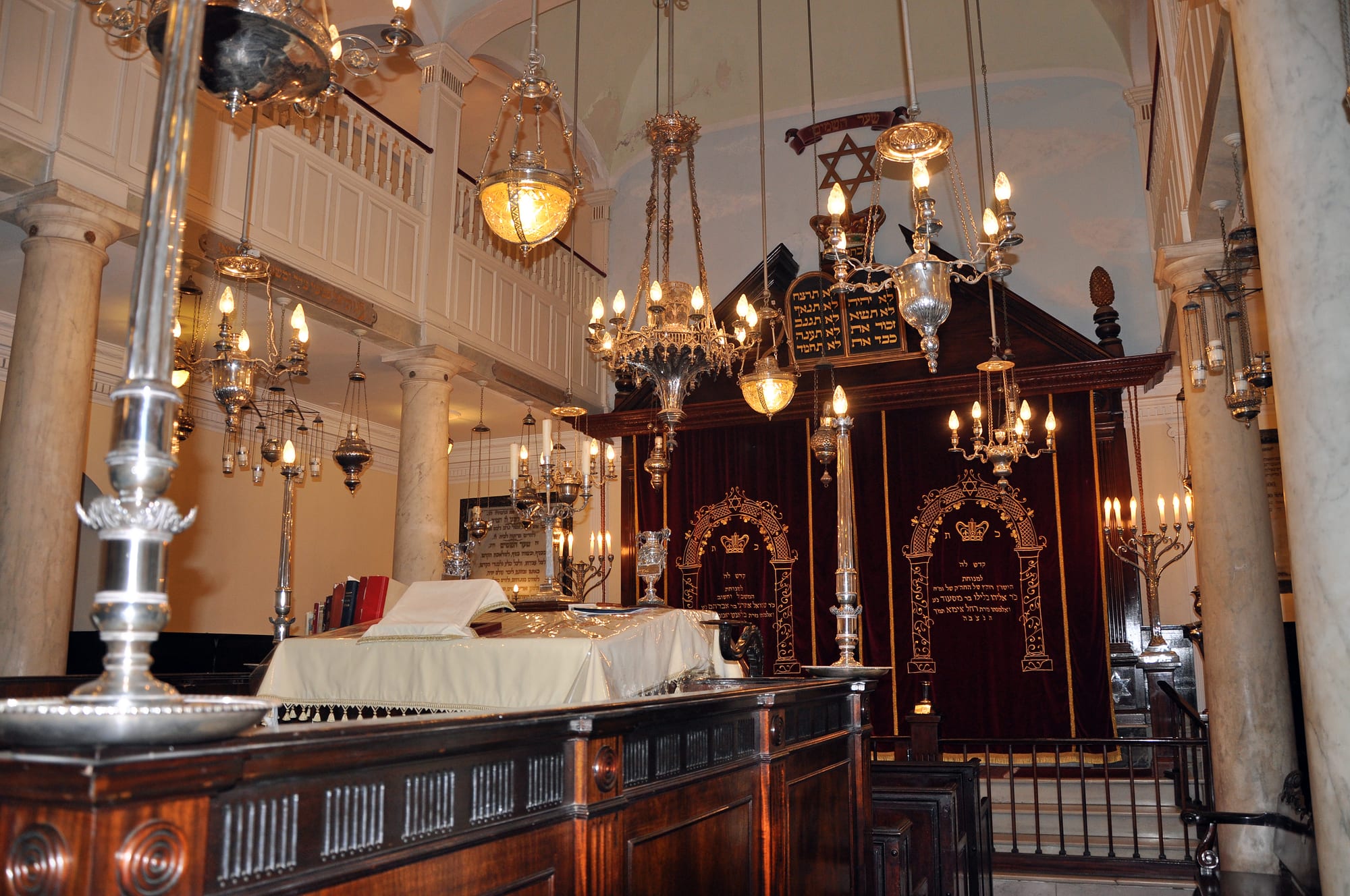 The interior of a synagogue with marble columns, a wooden gallery, silver hanging lamps with lit bulbs, a white-draped reading desk and a dark wooden ark with red and gold curtains