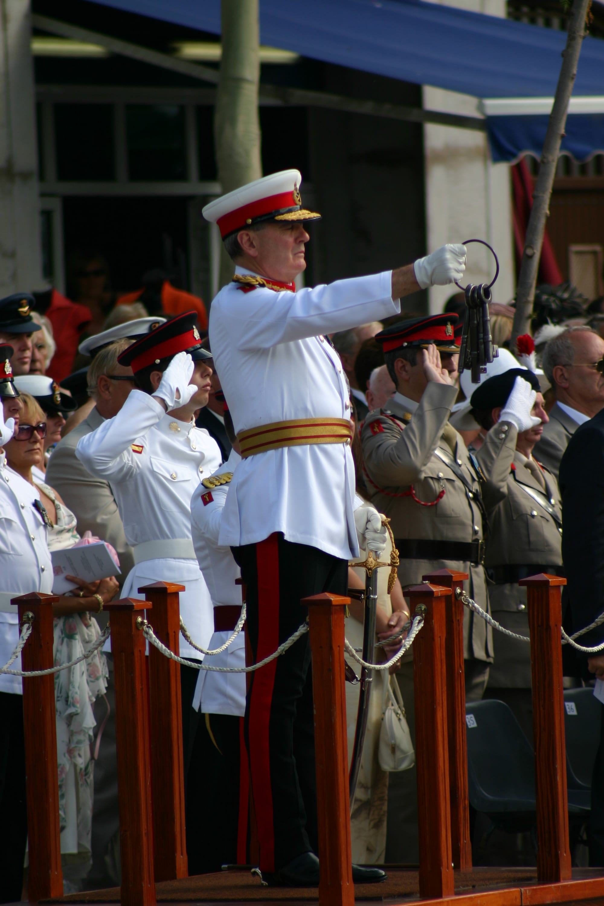 A man in a white ceremonial uniform with a gold sash and a red-banded peaked cap holds out a ring of large iron keys, while uniformed officers behind him salute
