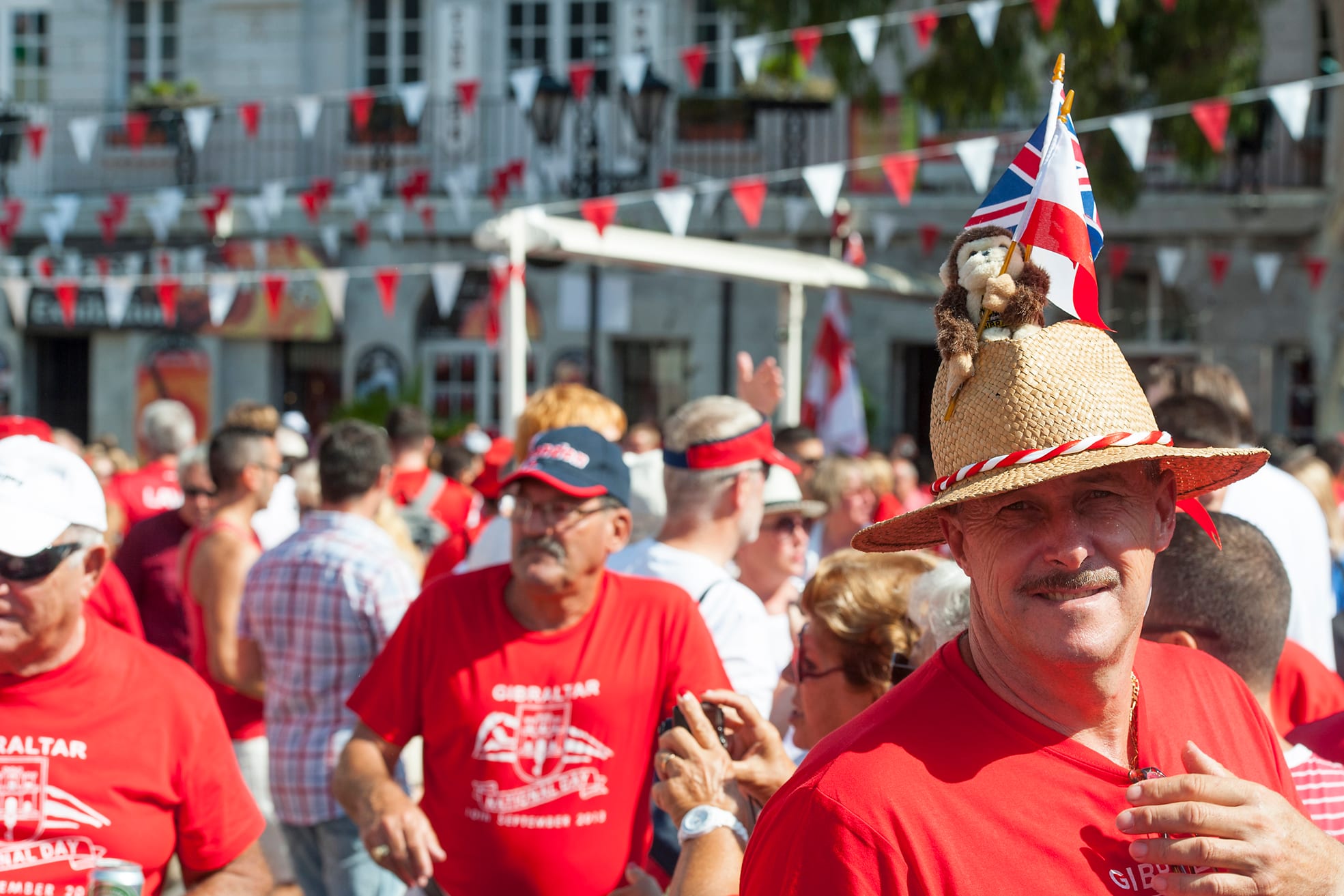 A crowd of people in red T-shirts in a square hung with red and white bunting, with a smiling man in the foreground wearing a straw hat topped with a toy monkey and a small Union flag