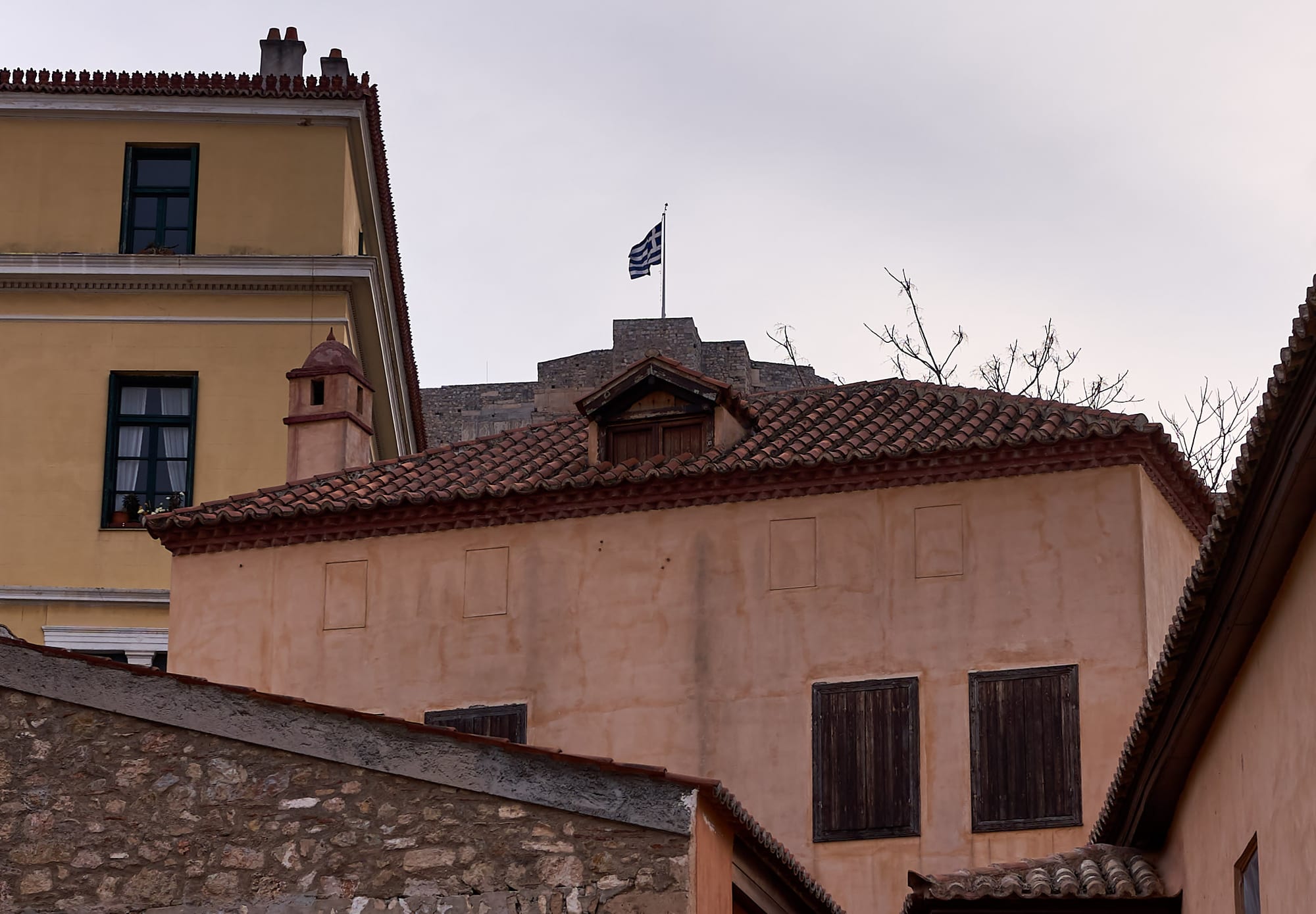 Old tiled-roof houses with shuttered windows in the Plaka district of Athens, a fortress wall visible behind