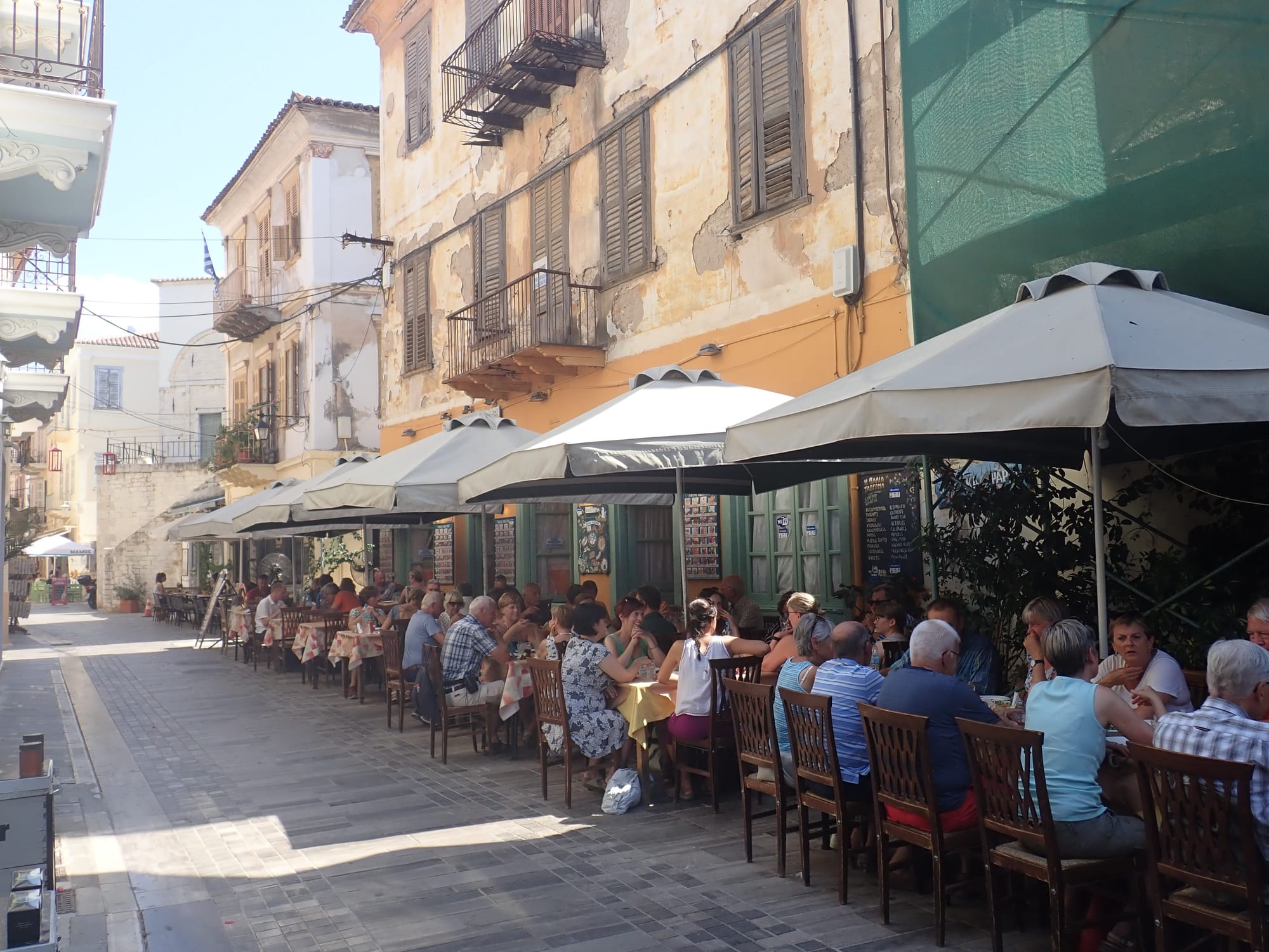 A row of outdoor taverna tables under awnings along a narrow street in Nafplio, filled with people eating lunch