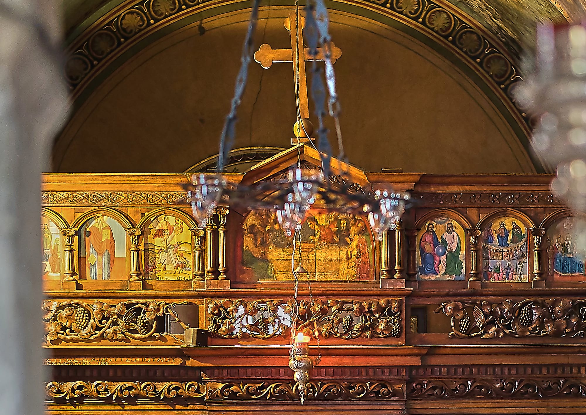 A carved and gilded wooden iconostasis with painted icons of saints and biblical scenes, topped by a wooden cross, with an oil lamp hanging in front
