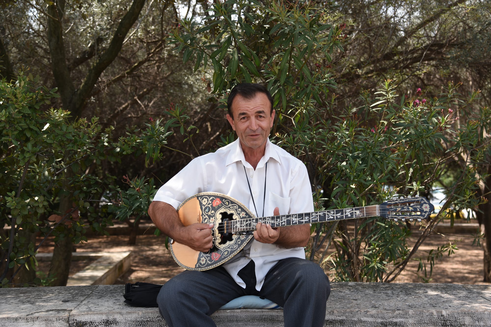 A man in a white shirt sits on a low stone wall playing a bouzouki with a decorated, inlaid body and a long neck, with trees behind him
