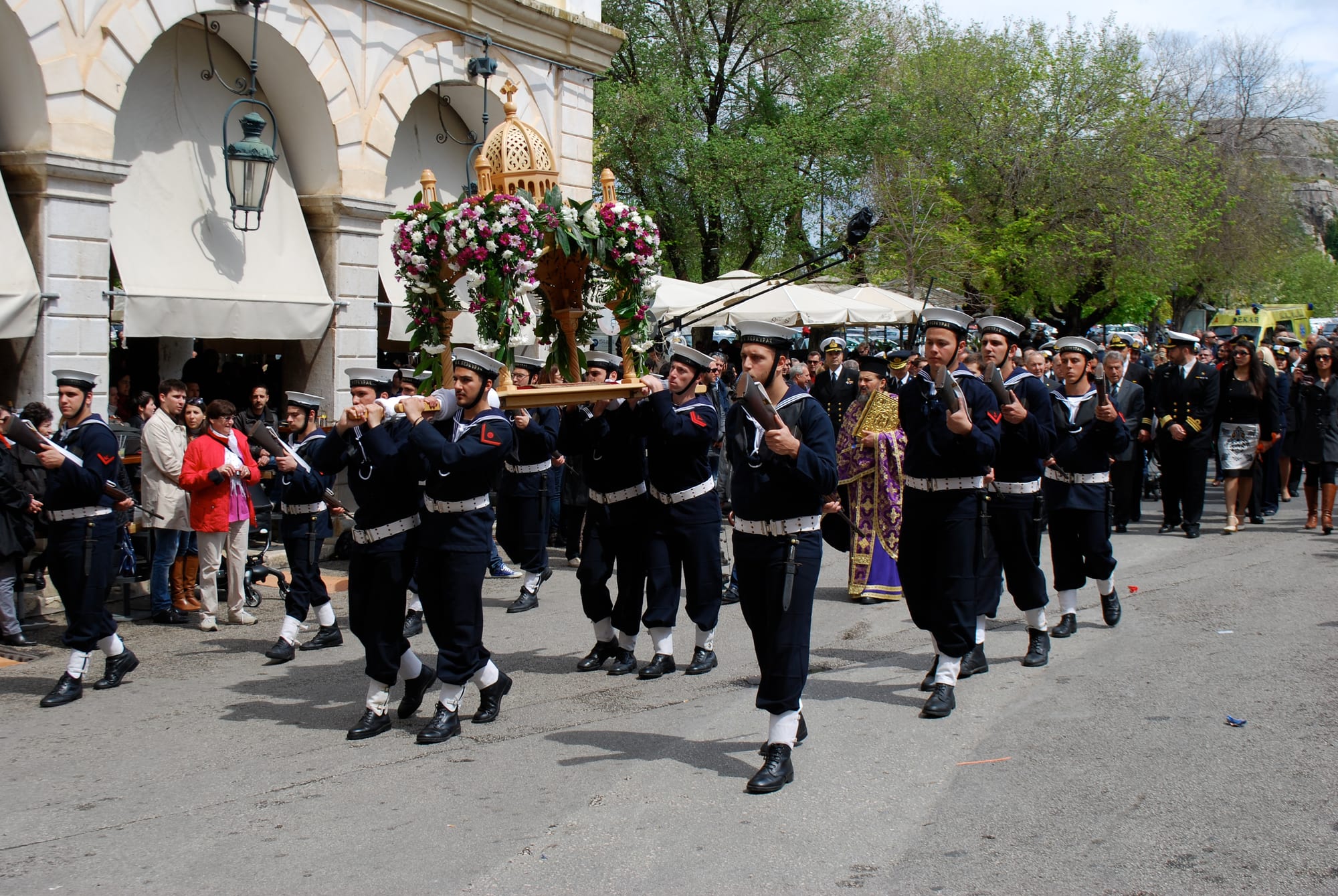 Sailors in dark uniforms and white caps carry a wooden bier covered in pink and white flowers along a street, followed by a priest in purple and gold vestments and a crowd
