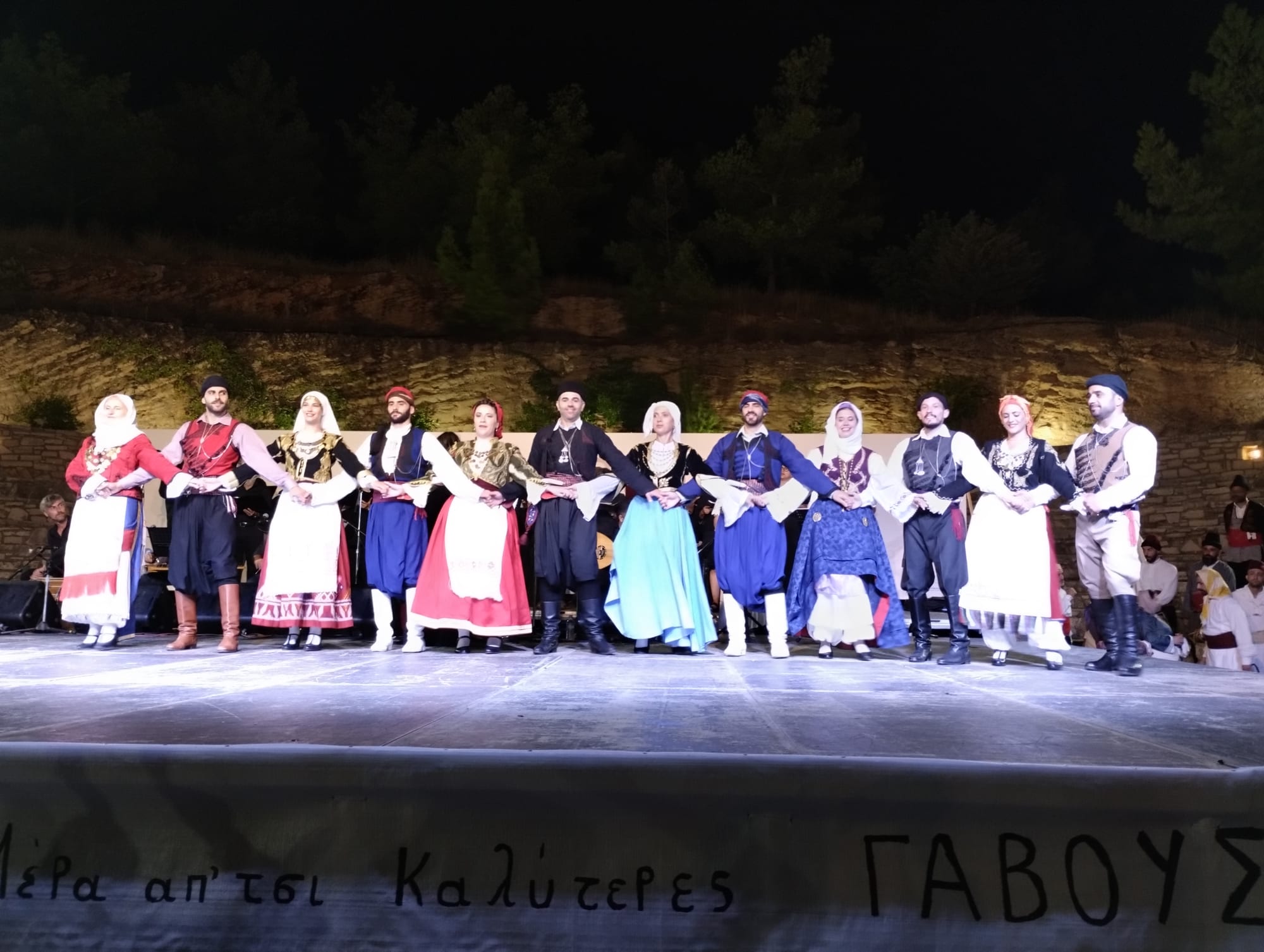A line of men and women in colourful traditional Cretan costumes hold hands on an open-air stage at night, with musicians behind them
