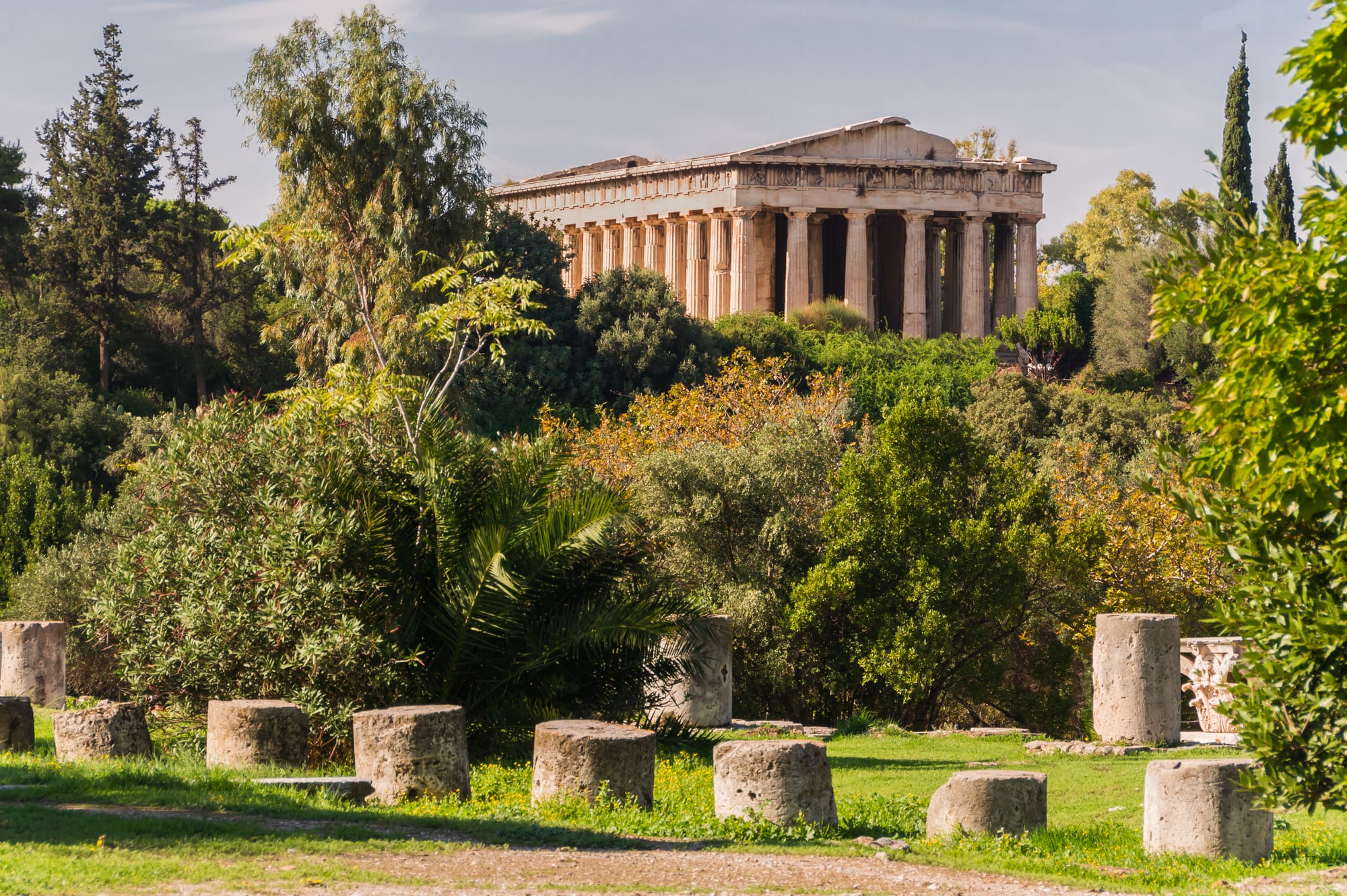 A Doric marble temple with its colonnade and pediment intact, standing on a wooded rise above the stumps of ancient columns