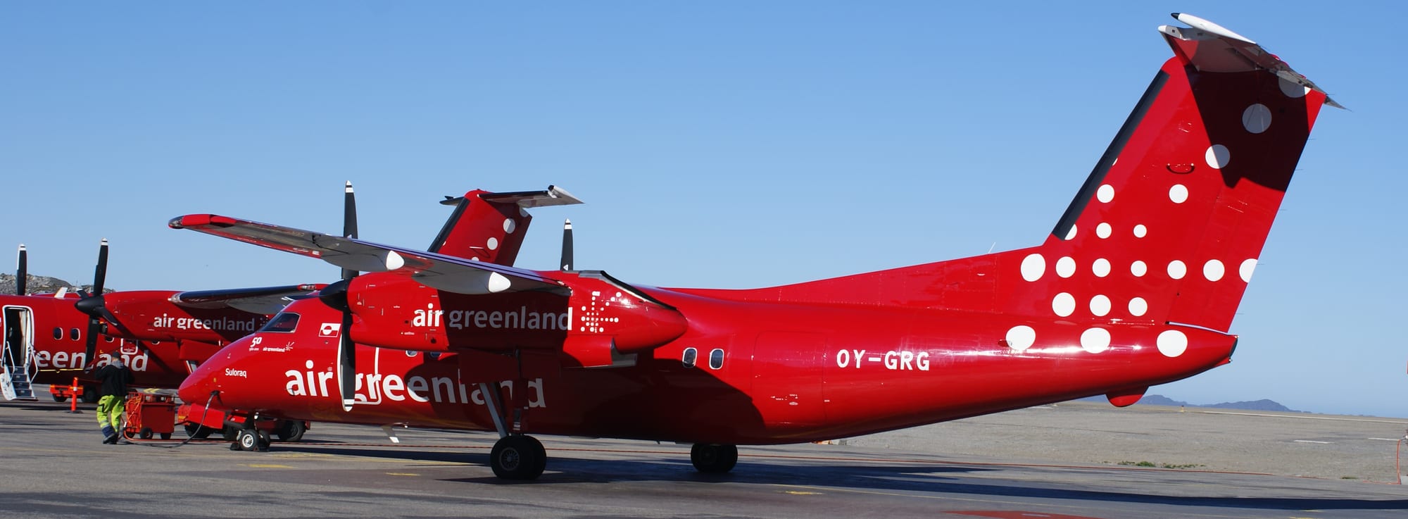 Two red Air Greenland Dash 8 propeller planes parked on the tarmac at Nuuk airport under a clear blue sky