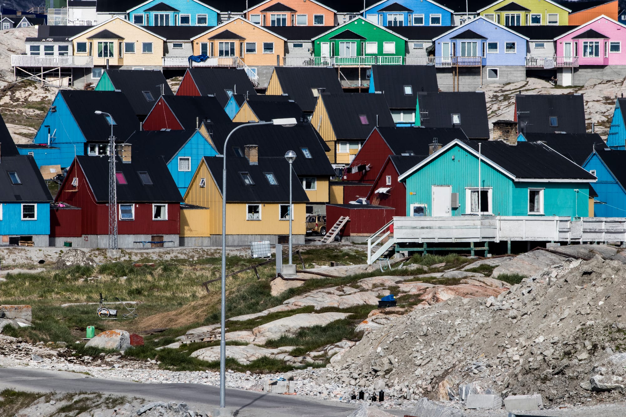 Rows of small red, blue, and yellow wooden houses on bare rock above a bay full of ice