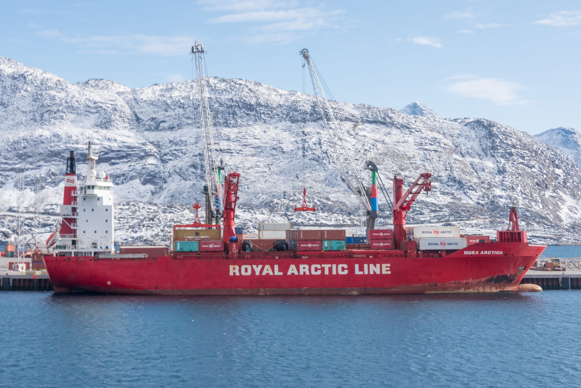 A container ship in a harbour below the low colourful buildings of Nuuk, snow-streaked mountains behind