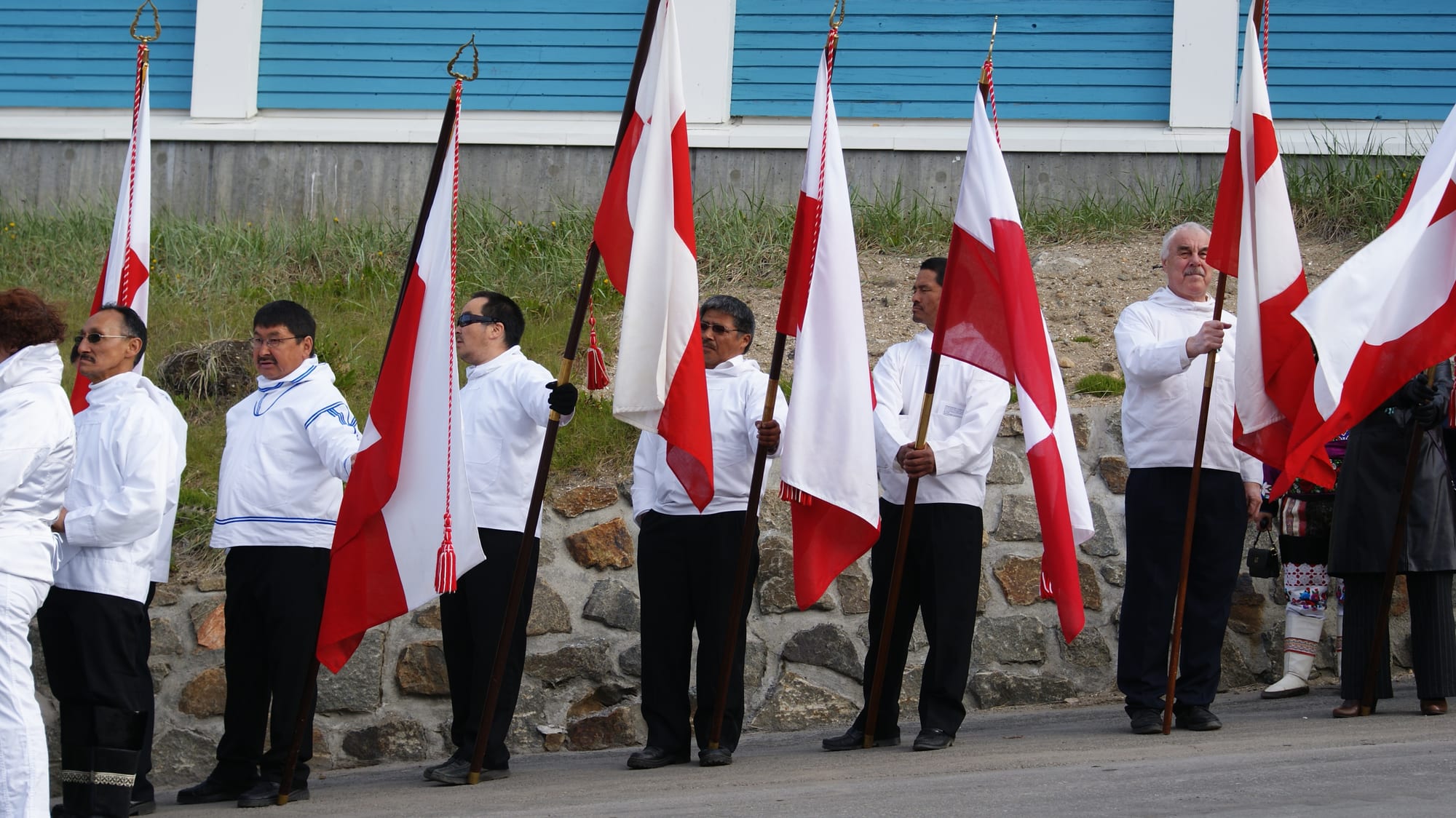 A row of men in white anoraks and dark trousers stand along a street holding tall poles with red and white Greenlandic flags, a pale blue wooden building behind them