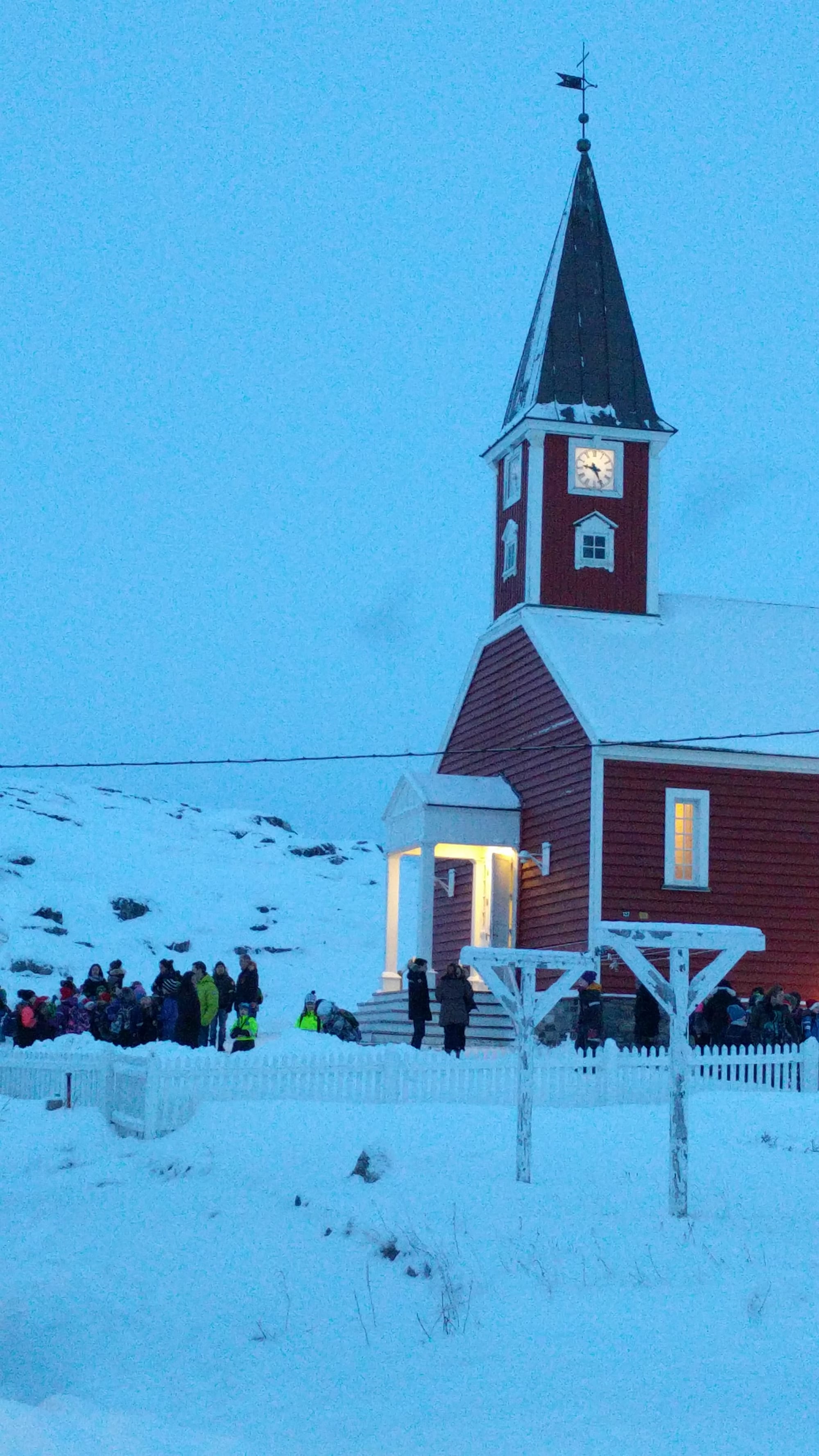 A red wooden church with a white-trimmed porch and a dark pointed spire stands in deep snow at dusk, its door lit, with groups of people in winter coats gathered outside