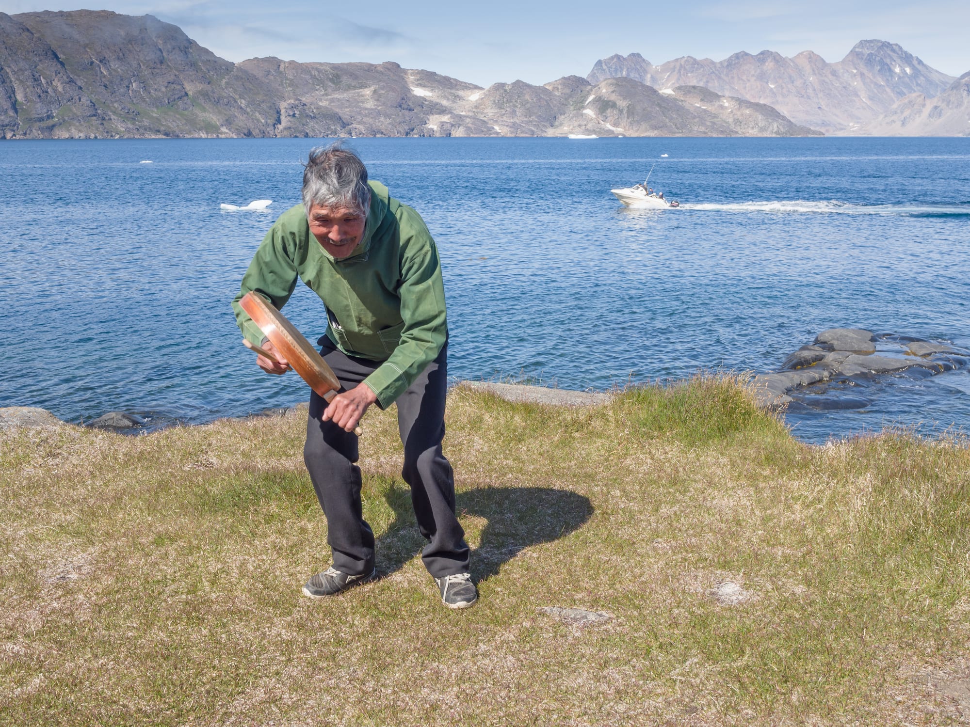 A grey-haired man in a green anorak bends forward on a grassy rise, striking the rim of a round frame drum with a stick, with a fjord, a small iceberg and mountains behind