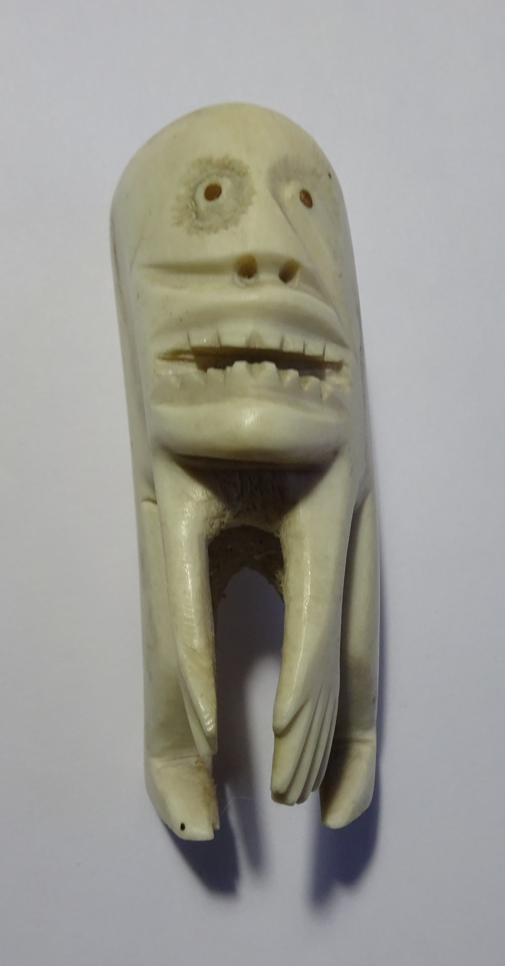 A small pale carved bone figure with a grimacing face, round eye holes, bared teeth and long clawed limbs, set against a white background