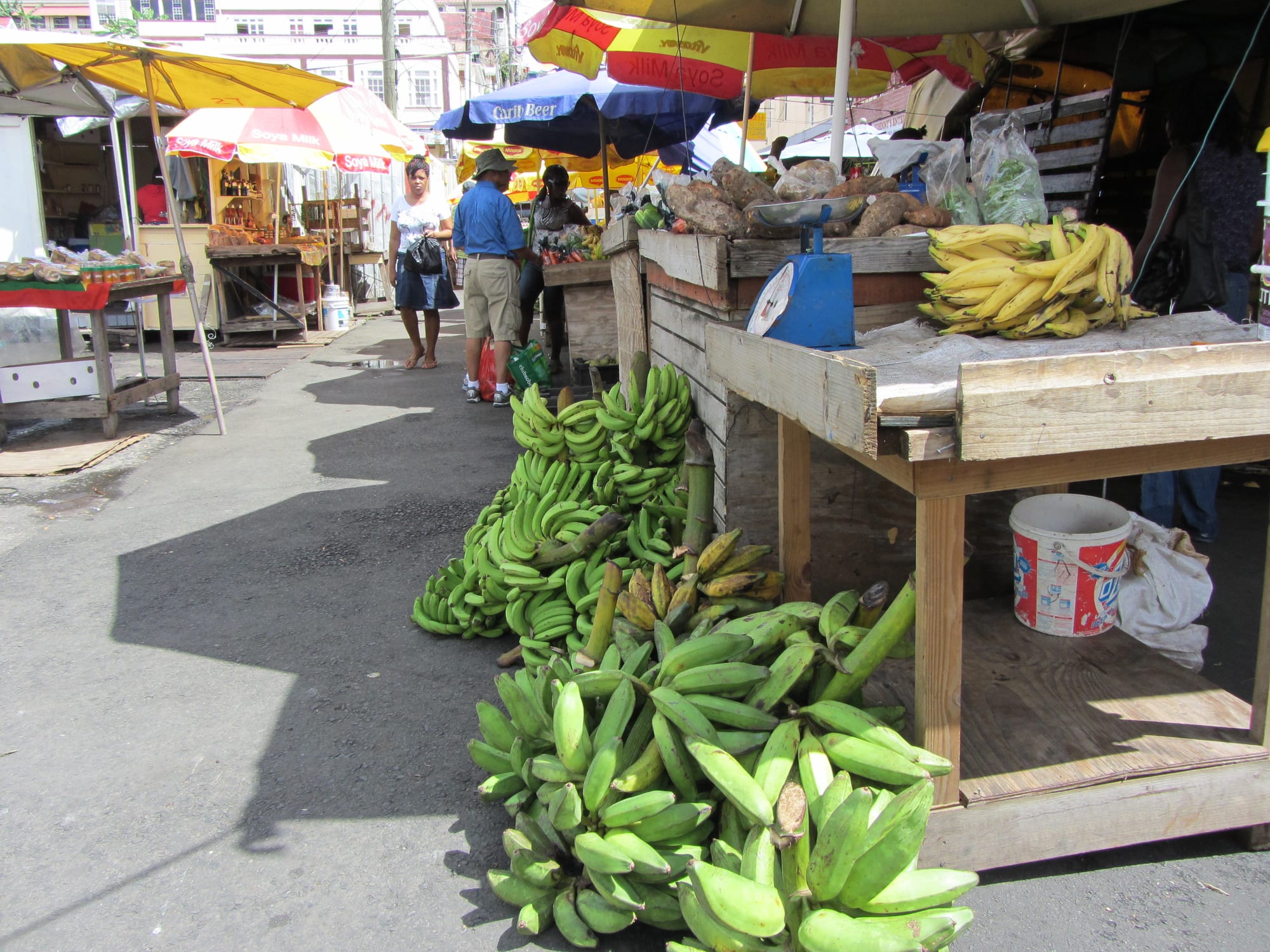 An outdoor market street in St George's with umbrella-covered stalls, bunches of green bananas, and shoppers walking past