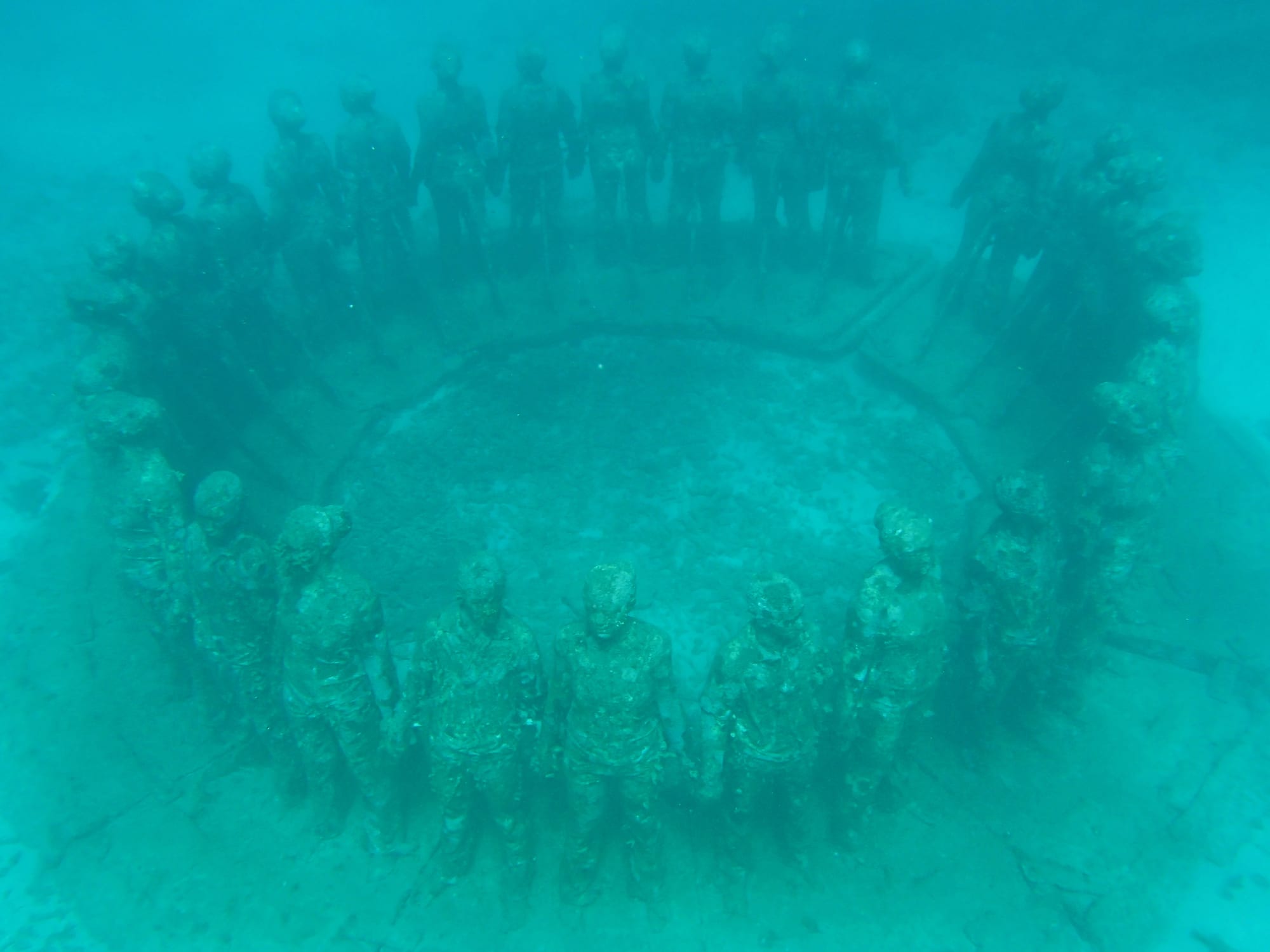 A ring of concrete human figures standing on the seabed, encrusted with coral