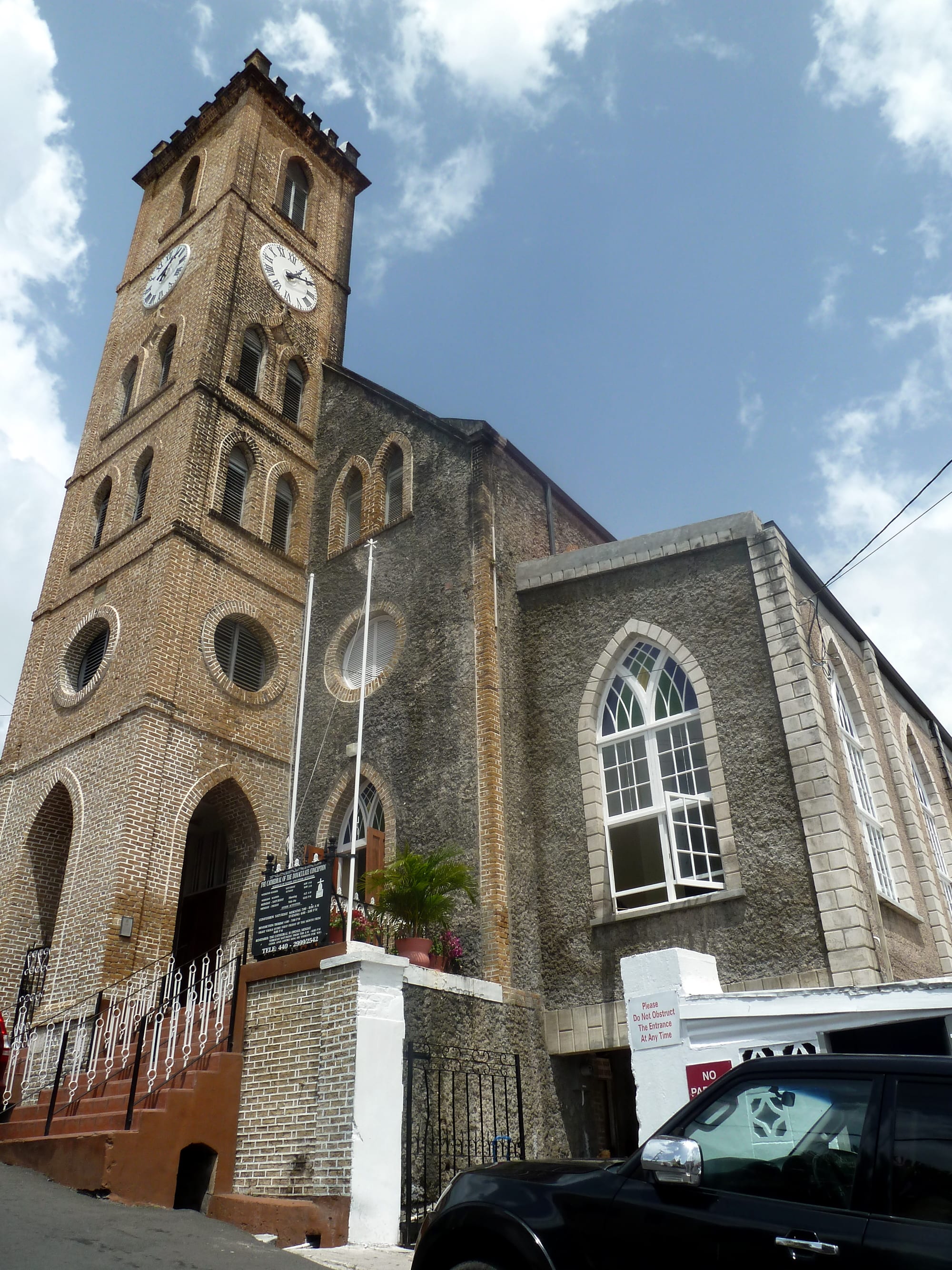 A tall brick bell tower with two clock faces and a crenellated top rises beside a grey stone church with arched windows, a noticeboard and potted plants by the entrance steps
