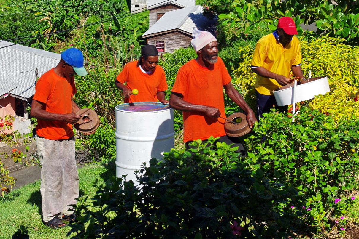 Four men in orange and yellow shirts and caps play steel pans of different sizes outdoors among flowering shrubs, with wooden houses on the slope behind