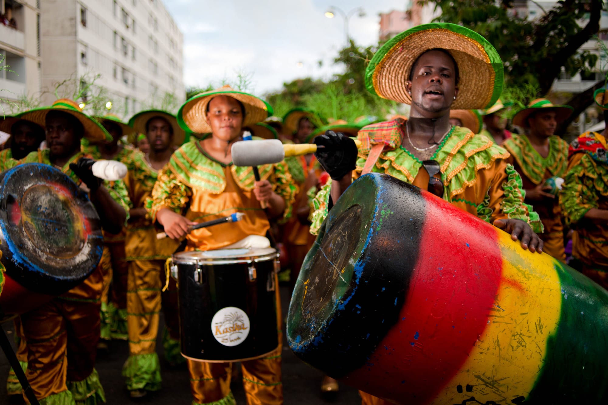 Carnival drummers in wide straw hats and matching gold and green ruffled costumes march in a street parade, one in front beating a large drum painted in red, yellow and green stripes