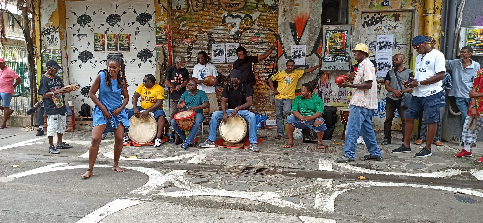 A woman in a blue dress dances barefoot on a paved street in front of a row of seated men playing hand drums, with a small crowd standing along a wall covered in posters and graffiti