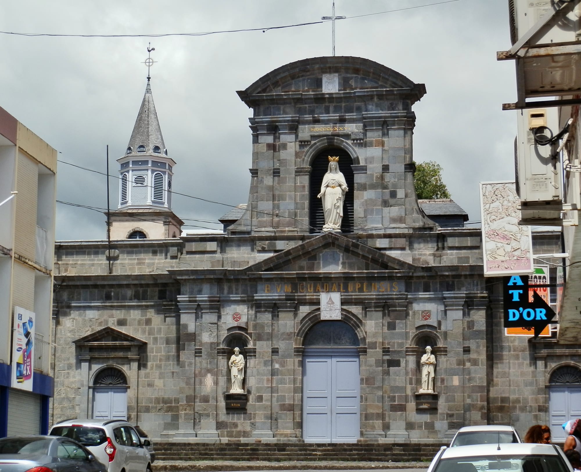 The grey stone facade of a church with a triangular pediment, statues in niches either side of a pale blue door, a crowned white statue of the Virgin in an arch above, and a slate-roofed bell tower to the left