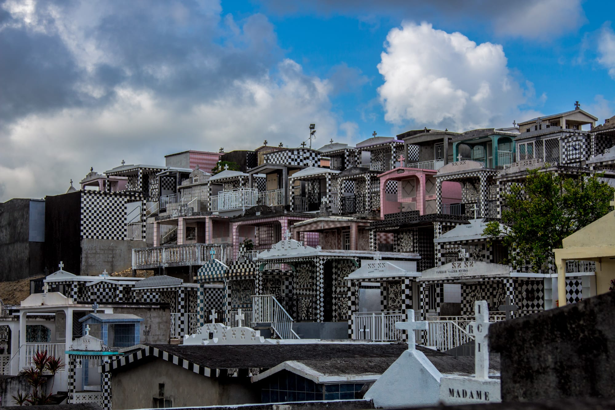 Tiers of family tombs covered in black and white checkerboard tiles, some with pink or white walls, small crosses and railings, climbing a hillside under a blue sky with white clouds