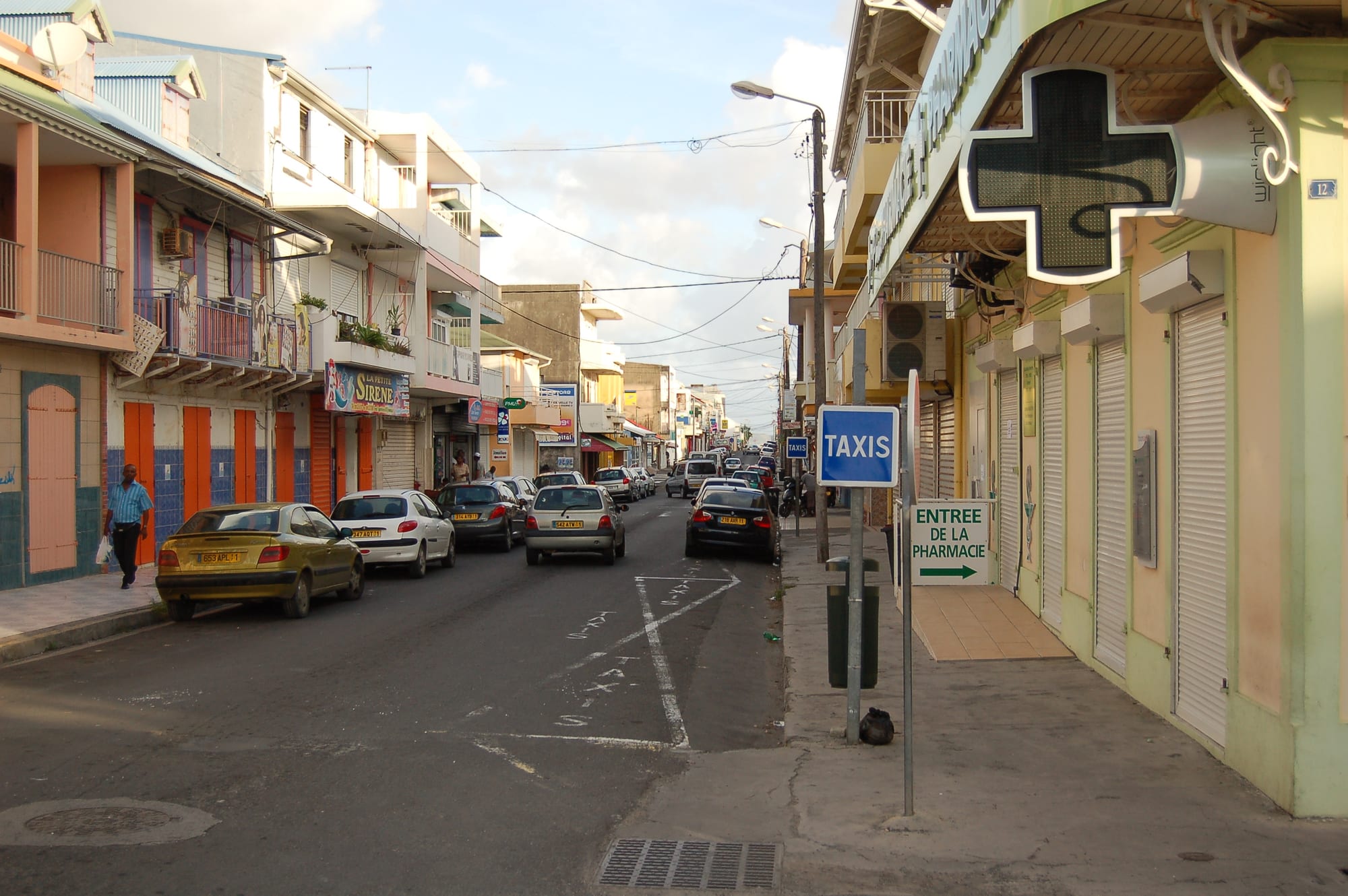 A street of shuttered shopfronts and balconied buildings in Le Moule, Guadeloupe, with parked cars and a taxis sign