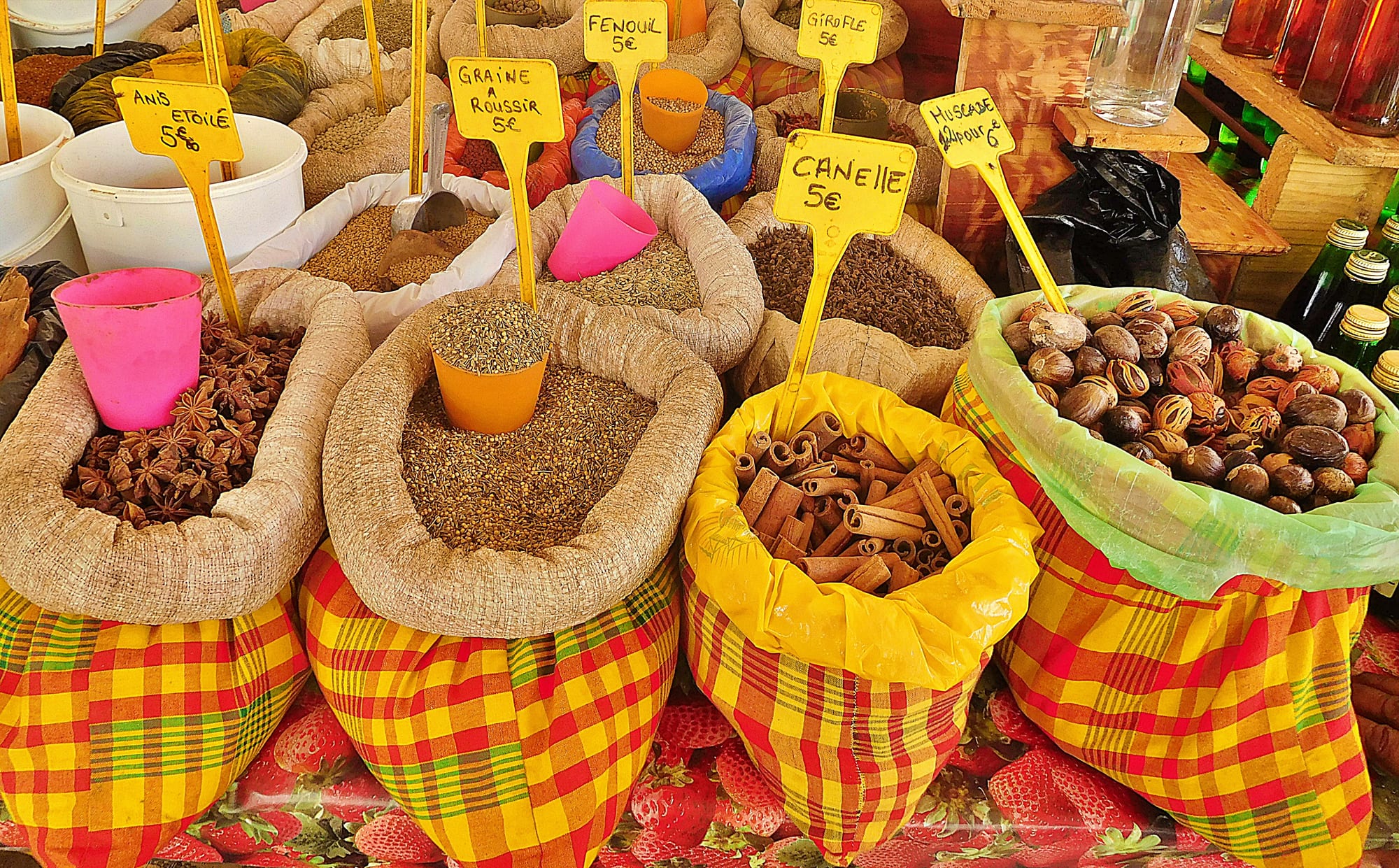 Burlap sacks of spices, wrapped in madras fabric and marked with hand-lettered price signs, on display at a market stall
