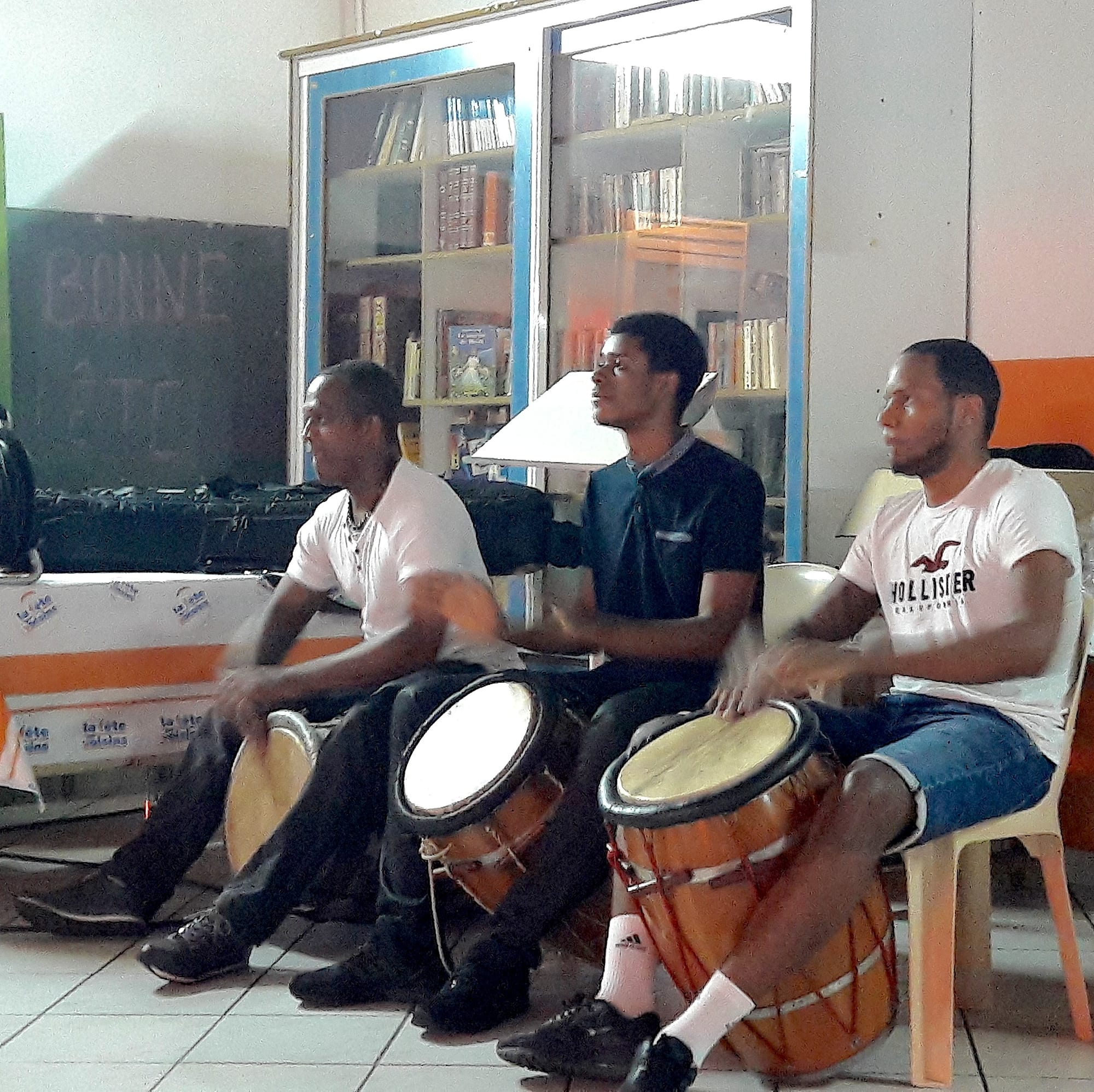 Three men playing traditional ka drums held between their knees at an indoor neighbourhood gathering