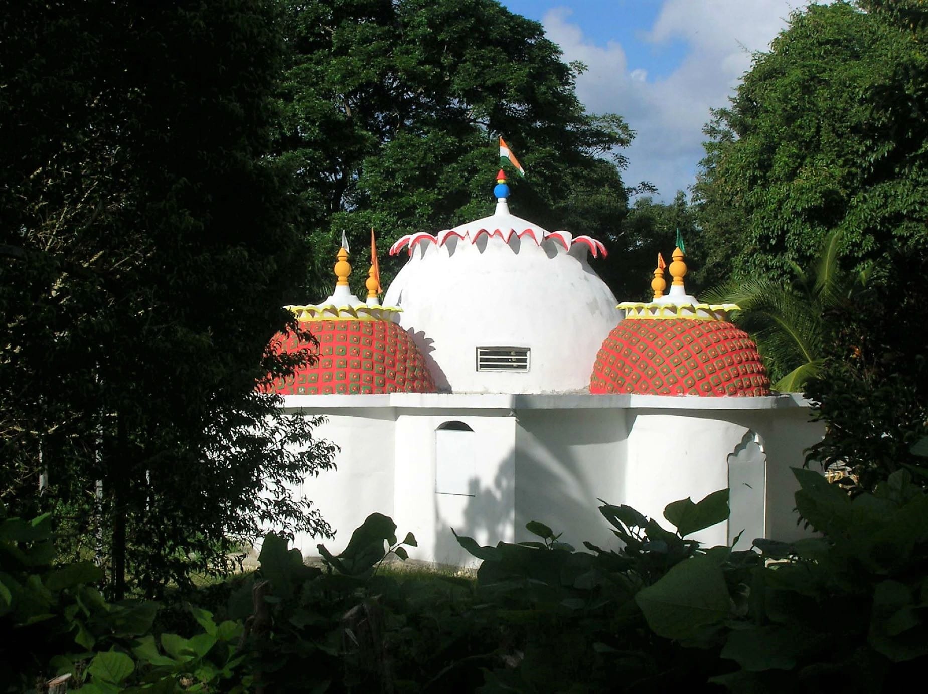 A small whitewashed temple with a white central dome and two red patterned side domes topped with gold finials, set among dense green trees