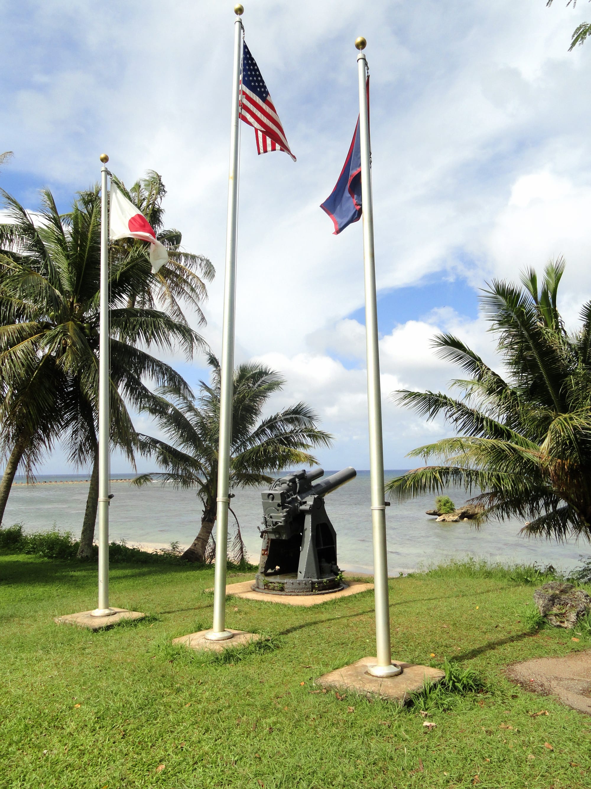US and Guam flags on poles beside a coastal battlefield memorial and old gun position